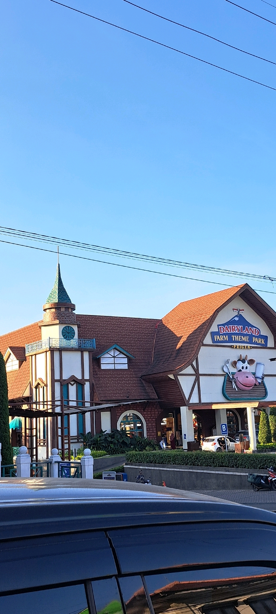 The image shows the entrance to Dairyland Farm Theme Park in Prigen, Indonesia. The building features a distinctive architecture with a steep, red-tiled roof, a tall tower with a green spire, and white and cream-colored walls. A large sign above the entrance reads "DAIRYLAND FARM THEME PARK" with an image of a smiling cow holding a fork and knife. Several cars and motorcycles are visible in the parking area in front of the park. The sky is clear and blue, indicating a sunny day. The overall scene depicts a tourist attraction in a well-maintained condition.