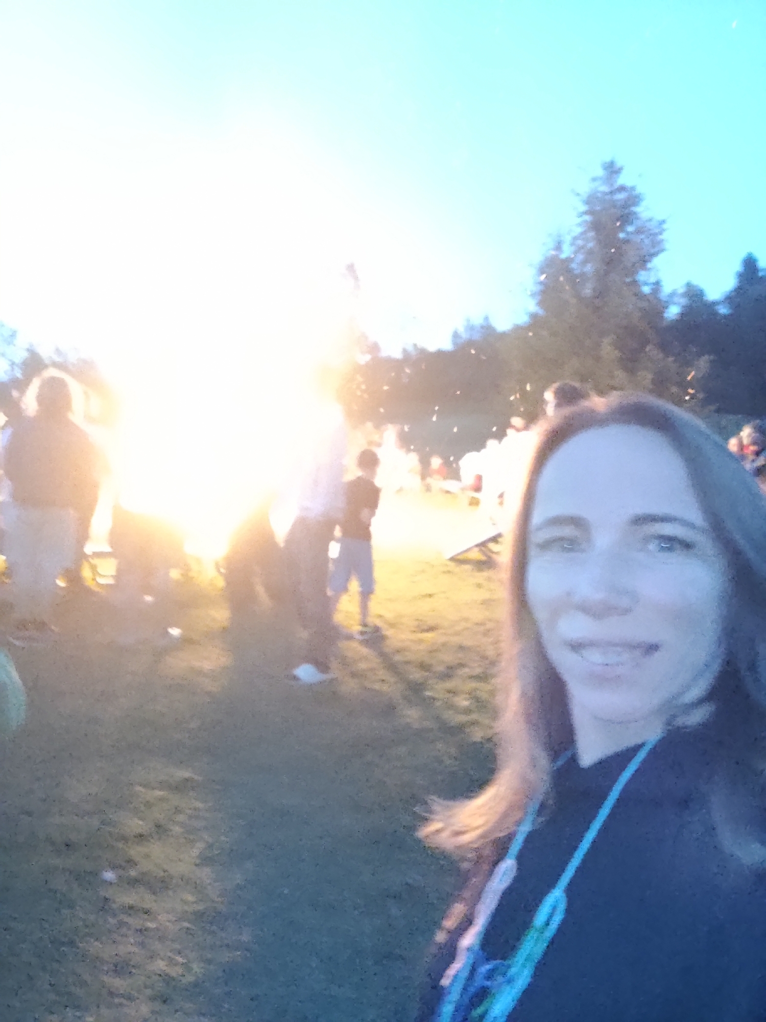 In the foreground, a woman with brown hair and blue eyes smiles at the camera. She is wearing a dark top and a colorful lanyard around her neck. Behind her, a large bonfire blazes, illuminating the scene. Several people, including children, are gathered around the fire, some appearing to be silhouetted against the flames. The environment is outdoors, with trees visible in the background and a grassy area in the foreground. The sky is a bright blue, indicating it is either dawn or dusk.