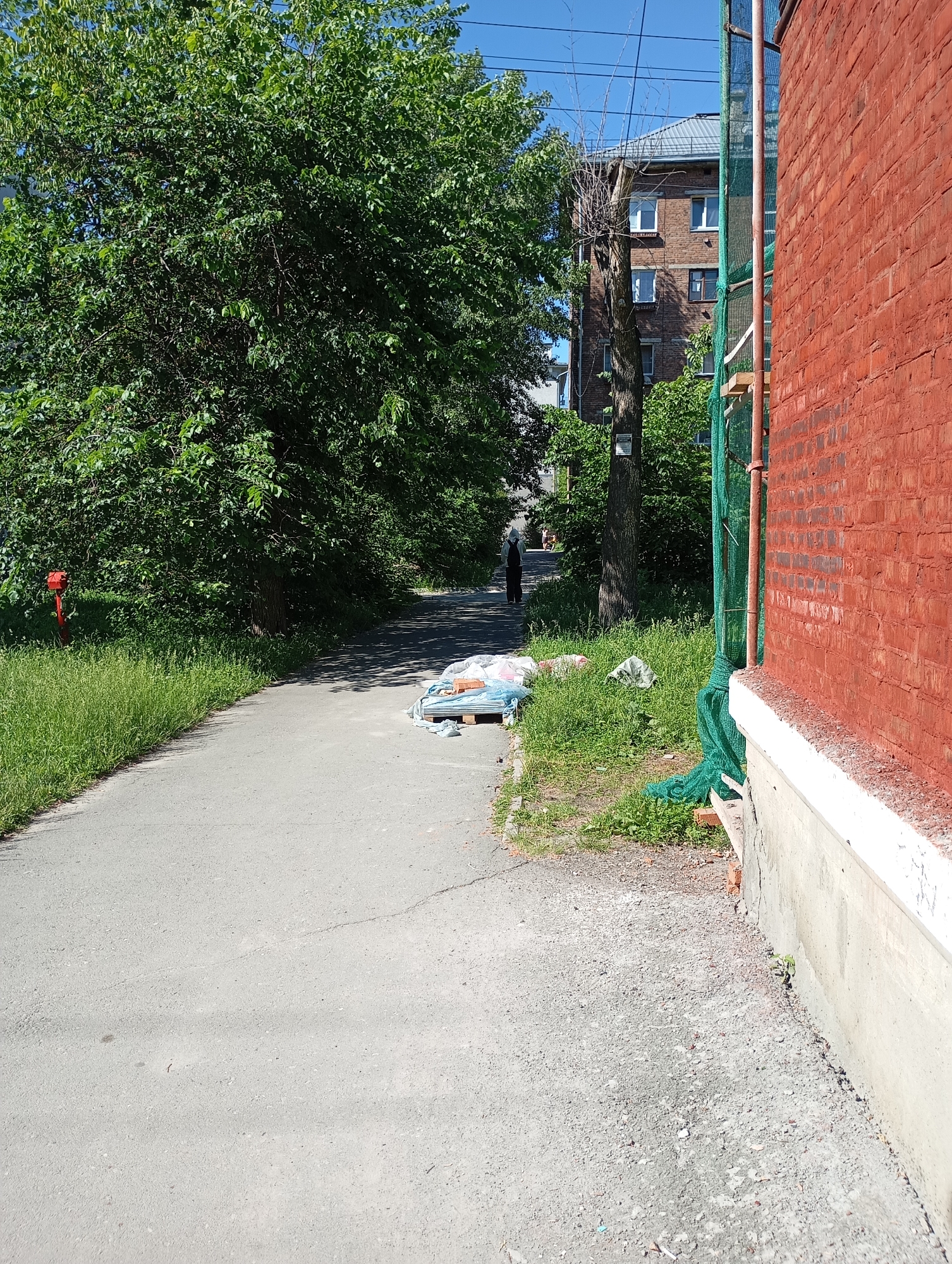 The image depicts an outdoor scene in Novosibirsk, Russia, on a sunny day. A paved pathway runs through the center of the frame, bordered by lush green grass on the left and a brick building on the right.
On the left side of the pathway, a vibrant green tree dominates the scene, its leaves casting dappled shadows. Near the tree, a red fire hydrant stands tall. Further down the path, a lone figure, dressed in dark clothing and a hood, walks away from the camera.
On the right side of the pathway, a portion of a red brick building is visible, with scaffolding and green netting surrounding it. The building appears to be under renovation or construction. In front of the building, a pile of construction materials, including plastic-wrapped items and wooden pallets, is stacked on the grass.
The ground is a mix of asphalt and gravel. The sky above is a clear, bright blue. The overall impression is of an urban environment, possibly a residential area, with evidence of ongoing construction work.
