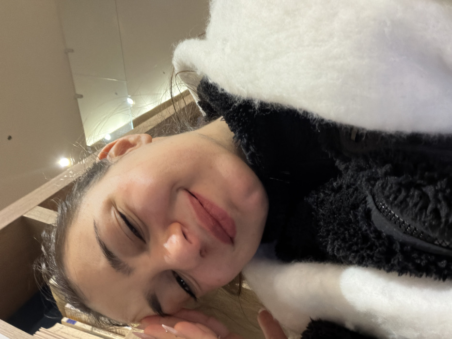 A young woman takes a selfie with a gentle smile. She is wearing a black and white fuzzy jacket. Part of a bookcase is visible in the left side of the frame, along with a ceiling with several lights above. Her nails are manicured, and she appears relaxed.