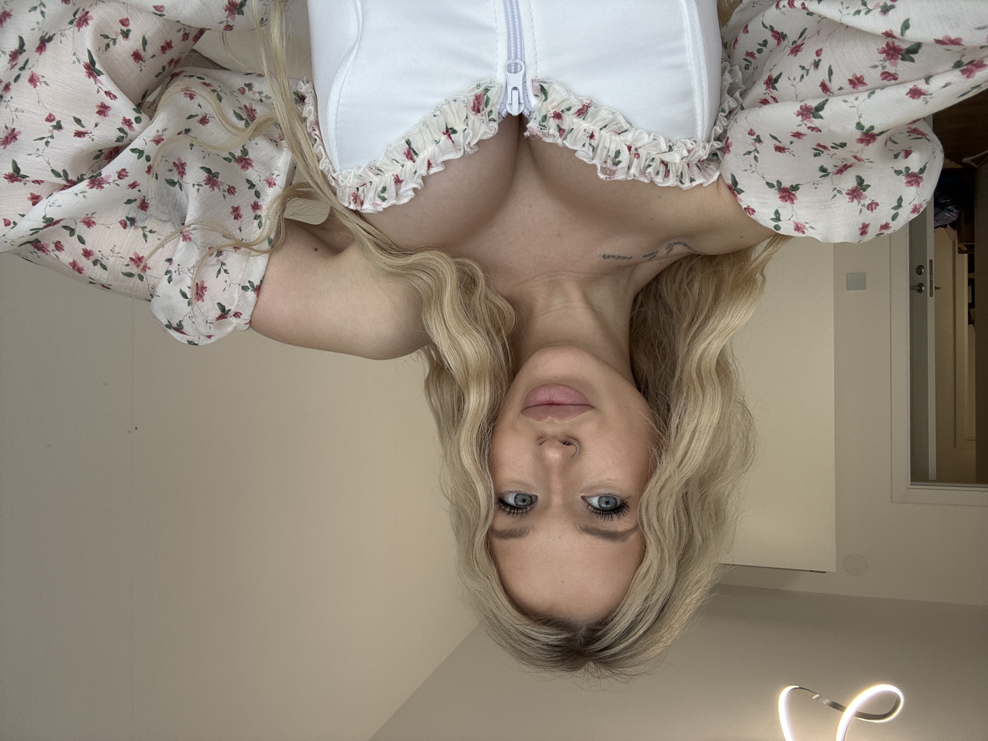 This is a vertically oriented, upside-down selfie of a young Caucasian woman with blonde hair and blue eyes. She is wearing a white corset-style top with a zipper and a ruffled trim, and a sheer white blouse with a pink floral pattern. Her hair is long and wavy, framing her face. She has light makeup on, and a nose ring. A tattoo is visible on her collarbone.
The background appears to be an indoor setting, possibly a room in an apartment. The lighting is soft and warm, suggesting it might be evening or an overcast day. The wall behind her is a neutral beige color, and a door and a window are visible to her right. A thin, illuminated light fixture is present in the lower right corner of the image.
The woman is looking directly at the camera with a neutral to slightly playful expression. The image is taken at a close-up to medium shot, focusing on her upper body and face. The overall mood is casual and personal, typical of a selfie. The context provided is Saint Petersburg, Russia. No specific text is visible in the image.