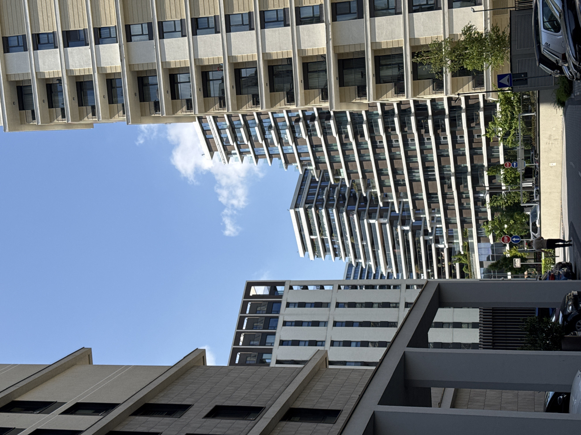 The image, taken in Belgrade, Serbia, presents an upward-looking view of the urban landscape, showcasing a variety of architectural styles under a bright, blue sky dotted with sparse white clouds.
Three distinct building types are visible:
1. A horizontally oriented building at the top of the image, characterized by its repetitively patterned facade with evenly spaced windows and balconies.
2. A tall, white building with a modern design, featuring rows of balconies.
3. A foreground building with a geometric, gray-toned facade, and a grid-like pattern of windows.
Visible at the right side of the image, a sidewalk is flanked by trees and street signs, including a stop sign and directional sign. A person is seen walking. Parked vehicles line the street.