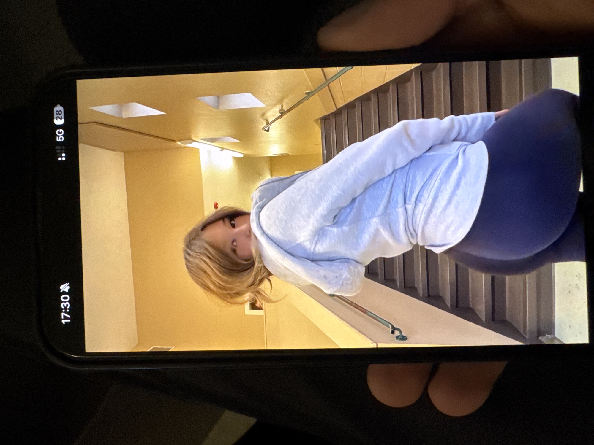 The image depicts a smartphone displaying a picture of a blonde woman wearing a white hoodie and blue leggings, standing on stairs inside a building with yellow walls and recessed ceiling lights. The phone screen shows the time as 17:30 and a 5G network connection. The phone is held by a person with tan skin. Location is indiscernible; Yerevan is not determinable.