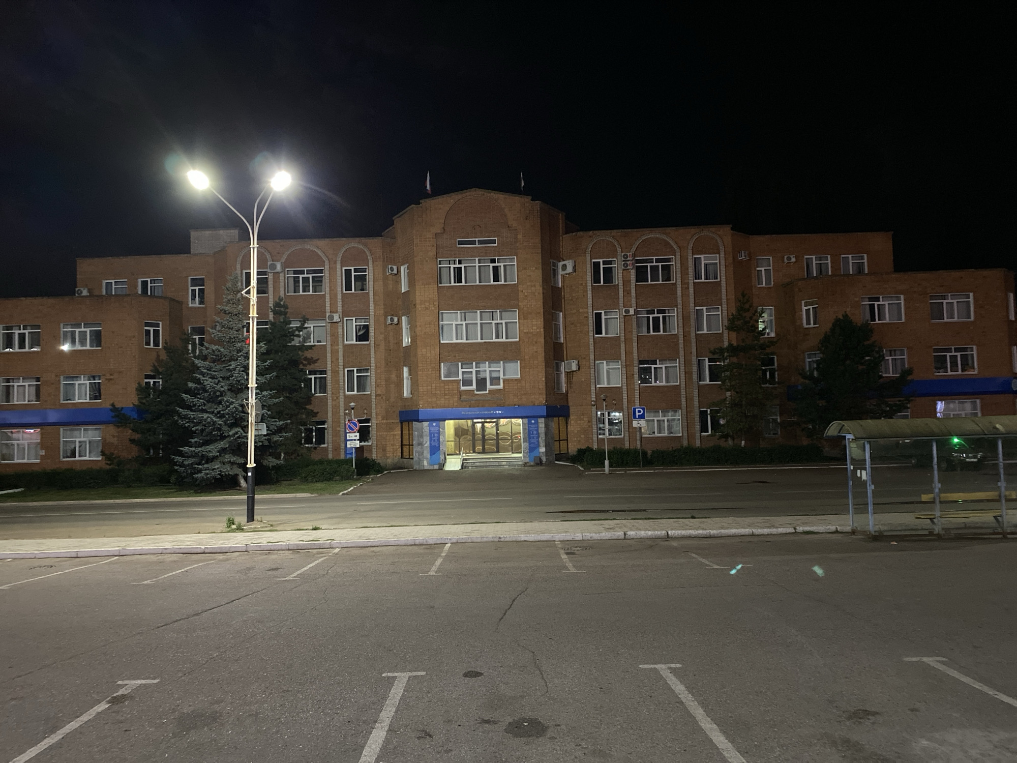 The image depicts a large, brick building at night, likely an administrative or institutional building, located in the city of Ishimbay, Russia. The building features multiple floors, numerous windows with white frames, and an arched entranceway with blue trim. A tall, double-headed streetlamp illuminates the foreground. In front of the building is a paved parking lot with white parking space lines. To the right, a bus stop shelter with a yellow bench is visible. The overall environment appears to be a street scene with a building and associated infrastructure. There are no people visible in the image.