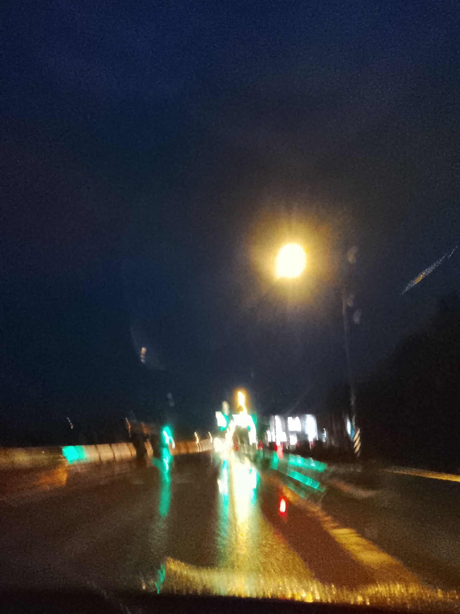 A blurred, low-resolution nighttime photograph captures a wet roadway, likely taken from inside a vehicle. A bright street lamp casts a strong glare in the upper center. The wet road reflects green, yellow, and red lights from other vehicles and roadside sources, creating elongated streaks of color. Building shapes with illuminated windows are visible in the background, indicating an urban or suburban environment. The sky is a dark, indistinct blue-black. Overall the scene suggests a rainy night drive in an unknown city.