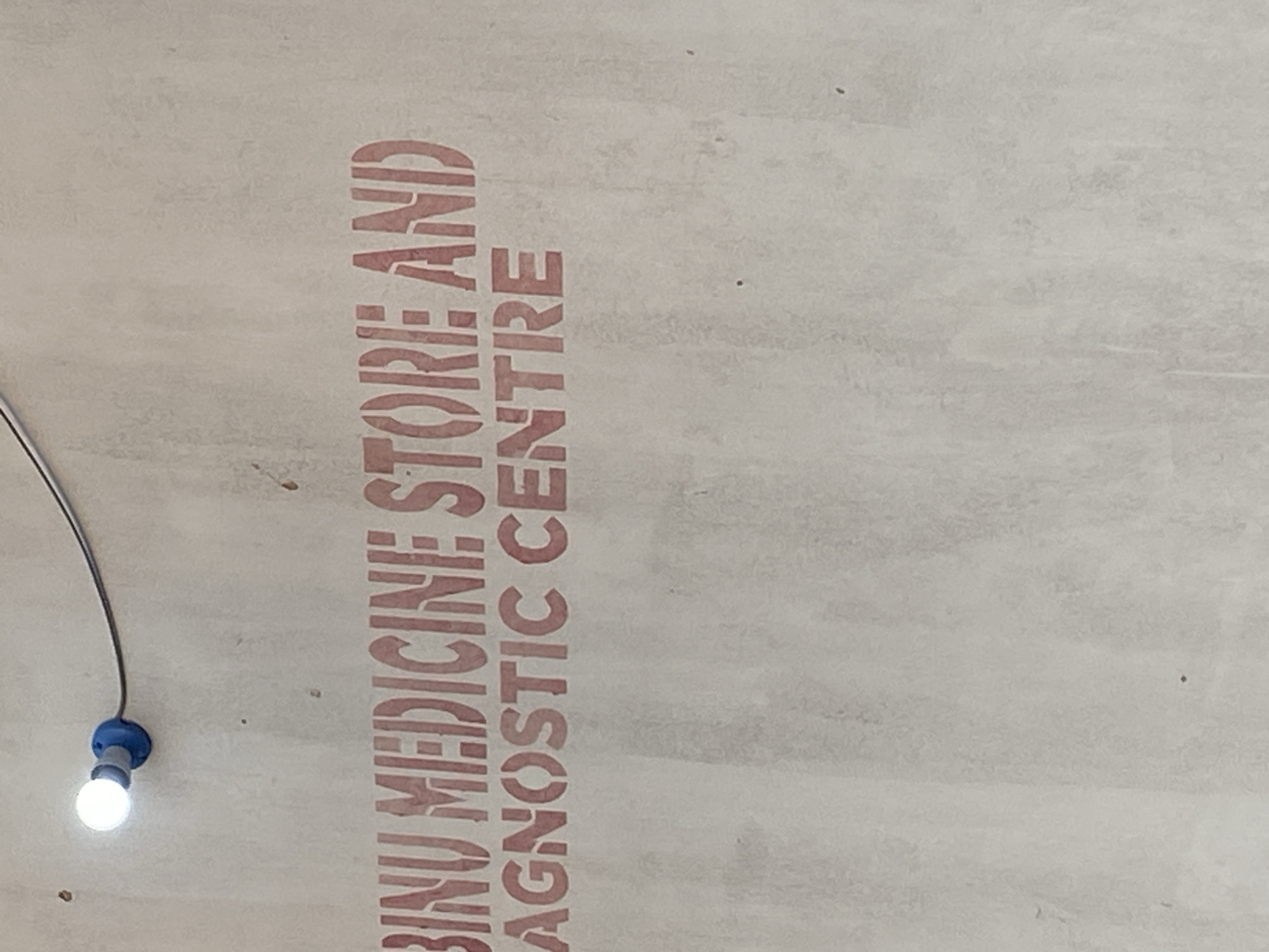 The image displays a red stencil painted on a textured off-white wall, likely indicating signage for a business. The text reads "BINU MEDICINE STORE AND DIAGNOSTIC CENTRE". A bright white LED light bulb is visible in the upper left corner of the frame, attached to a blue electrical fitting and connected to a dark wire. The wall exhibits a weathered appearance with some faint darker marks and smudges. The scene appears to be an exterior or interior wall in a public space, potentially a storefront. The location information "Jauro Yinu, Nigeria" is not visually represented in the image itself and must be inferred from external context.
