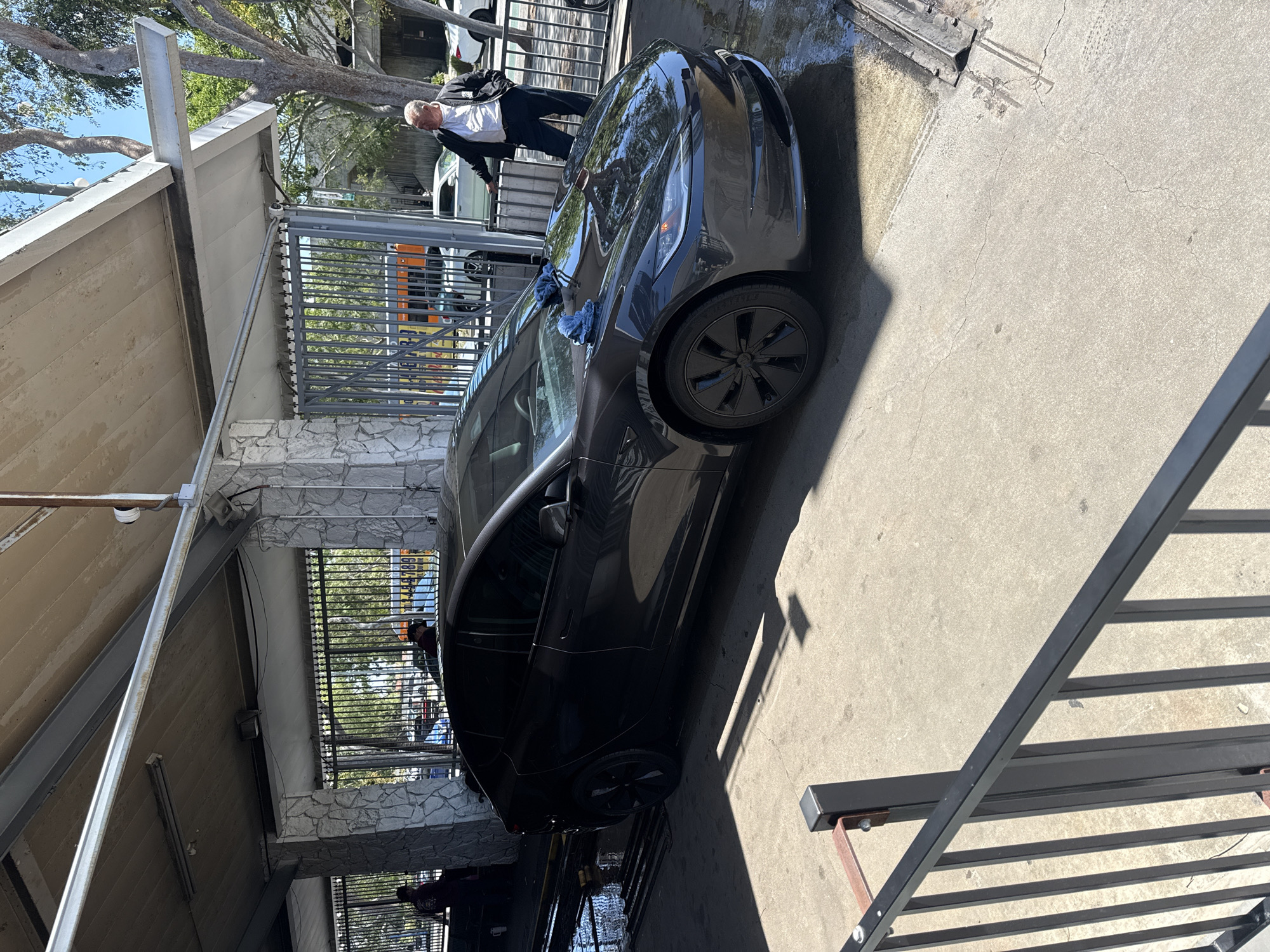 The image shows a dark-colored Tesla sedan parked in what appears to be a car wash or detailing location in West Hollywood, California. A man wearing a white shirt and dark pants is near the front of the car. The location is identifiable by the architectural details of the buildings and fences surrounding the vehicle. Water residue indicates the car has recently been washed. The setting appears to be an outdoor work area.