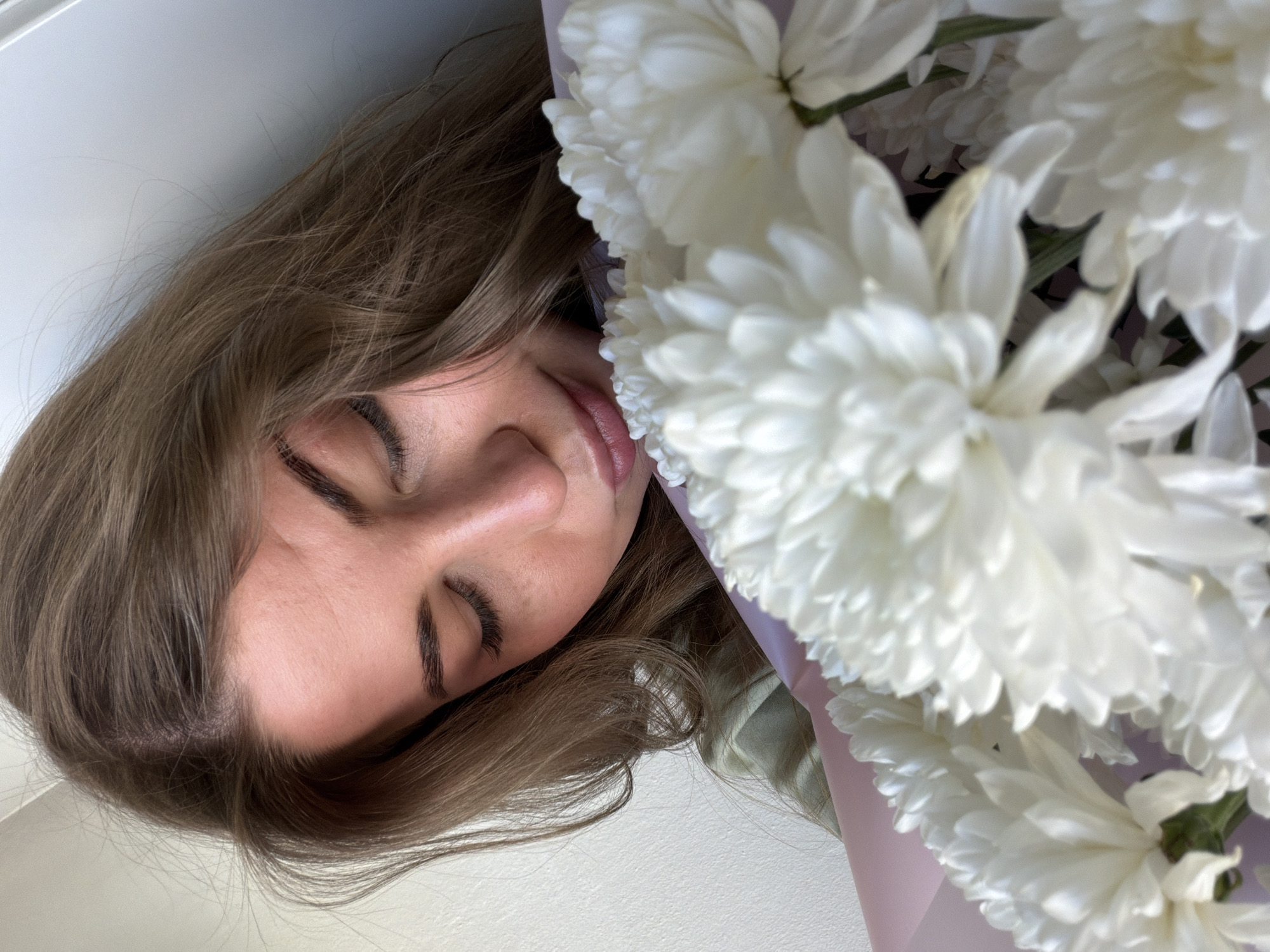 A close-up, slightly angled shot shows a woman's face partially obscured by a bouquet of white flowers. Her eyes are closed, and she has light brown hair. The background appears to be a plain white surface. There is no indication of the location being Chaikovsky, Russia in the image.