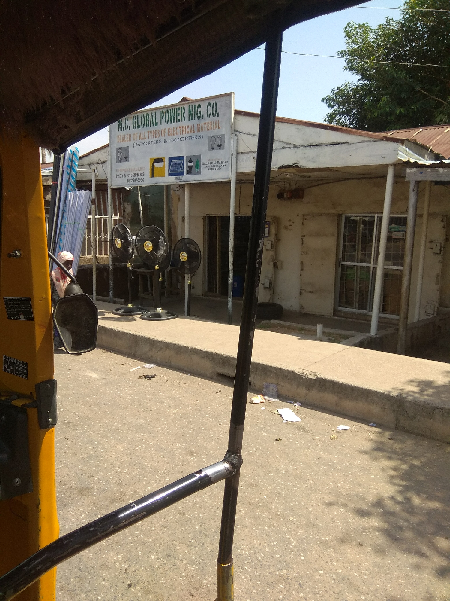 The image shows a street scene in Jalingo, Nigeria. In the foreground, the yellow body and black metal frame of a vehicle, possibly an auto-rickshaw, are visible, partially obscuring the view. A large black side mirror is attached to the vehicle's frame. A person wearing a light-colored headscarf and a patterned shirt stands behind the mirror. Beyond the vehicle, a building with a white facade and a corrugated iron roof is visible. A sign board above the building's entrance reads "M.C. GLOBAL POWER NIG. CO. DEALER OF ALL TYPES OF ELECTRICAL MATERIAL (IMPORTERS & EXPORTERS)." The sign also features various icons representing electrical items like light bulbs, solar panels, and a fan. Below the sign, contact information, including a phone number and address in Jalingo, Kano State, is displayed. In front of the building, three black oscillating fans on stands are placed on a concrete walkway. The ground is paved with asphalt, and there is some scattered debris on the road and walkway. Trees with green foliage are visible in the background against a clear blue sky. The overall impression is that of a commercial area on a sunny day.