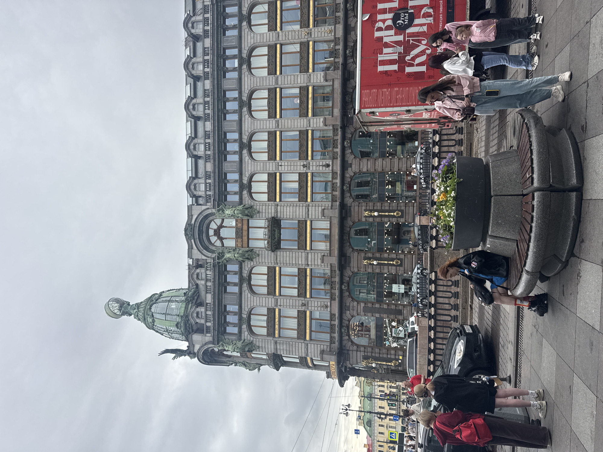 The image shows a wide shot of Singer House, also known as the Dom Knigi, located on Nevsky Prospekt in Saint Petersburg, Russia. The iconic building features a prominent green dome topped with a globe and an eagle. In the foreground, several people are gathered. On the left, three young women are standing near a black car. One is wearing a red backpack, another a black hoodie, and the third a red jacket. To the right, a young woman with red hair is sitting on a circular bench, wearing a blue dress and a black jacket. In the center, a large planter filled with flowers sits on the bench. Further to the right, a group of four people, three women and a young girl, are standing near a red truck with Russian text that appears to advertise a beverage company. The sky is overcast, and the scene takes place on a paved pedestrian area.