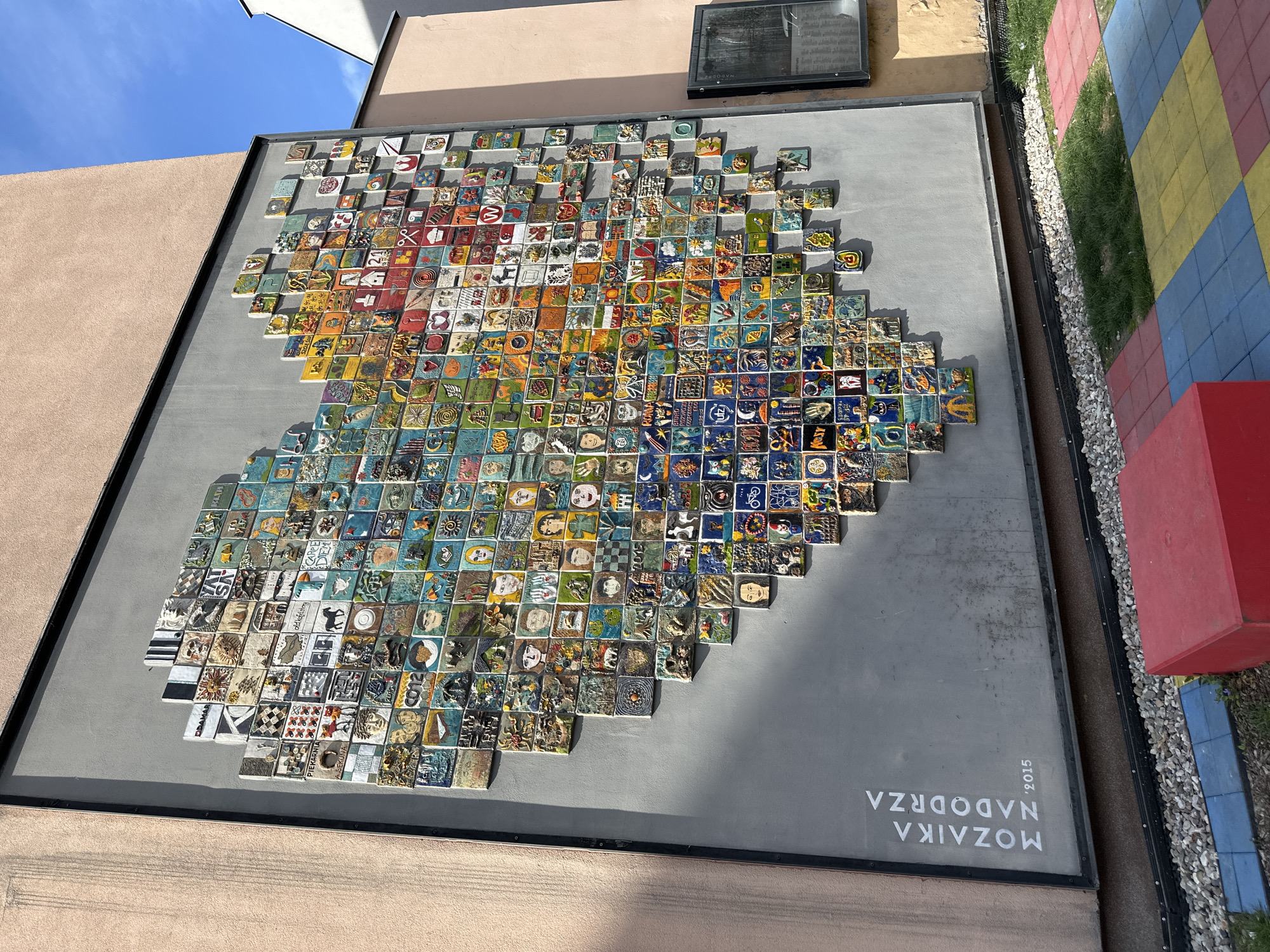 A large, square mosaic panel is affixed to a building's exterior wall. The mosaic features numerous square tiles, each individually decorated with various images and designs, arranged together to form a rough, organic shape. The overall color scheme is diverse, with reds, oranges, greens, blues, and whites prominently featured. The background behind the mosaic is gray.
Below the mosaic, in white lettering, are the words "MOZAIKA NADODRZA '2015". The panel is surrounded by a black frame.
To the right of the mosaic, a narrow strip of landscaping can be seen, with small plants, pebbles, and multi-colored paving stones. The building itself is a light tan color. The sky is visible in the upper left corner, with some blue visible. The building is the "Nadodrze Mosaic" in Wrocław, Poland.