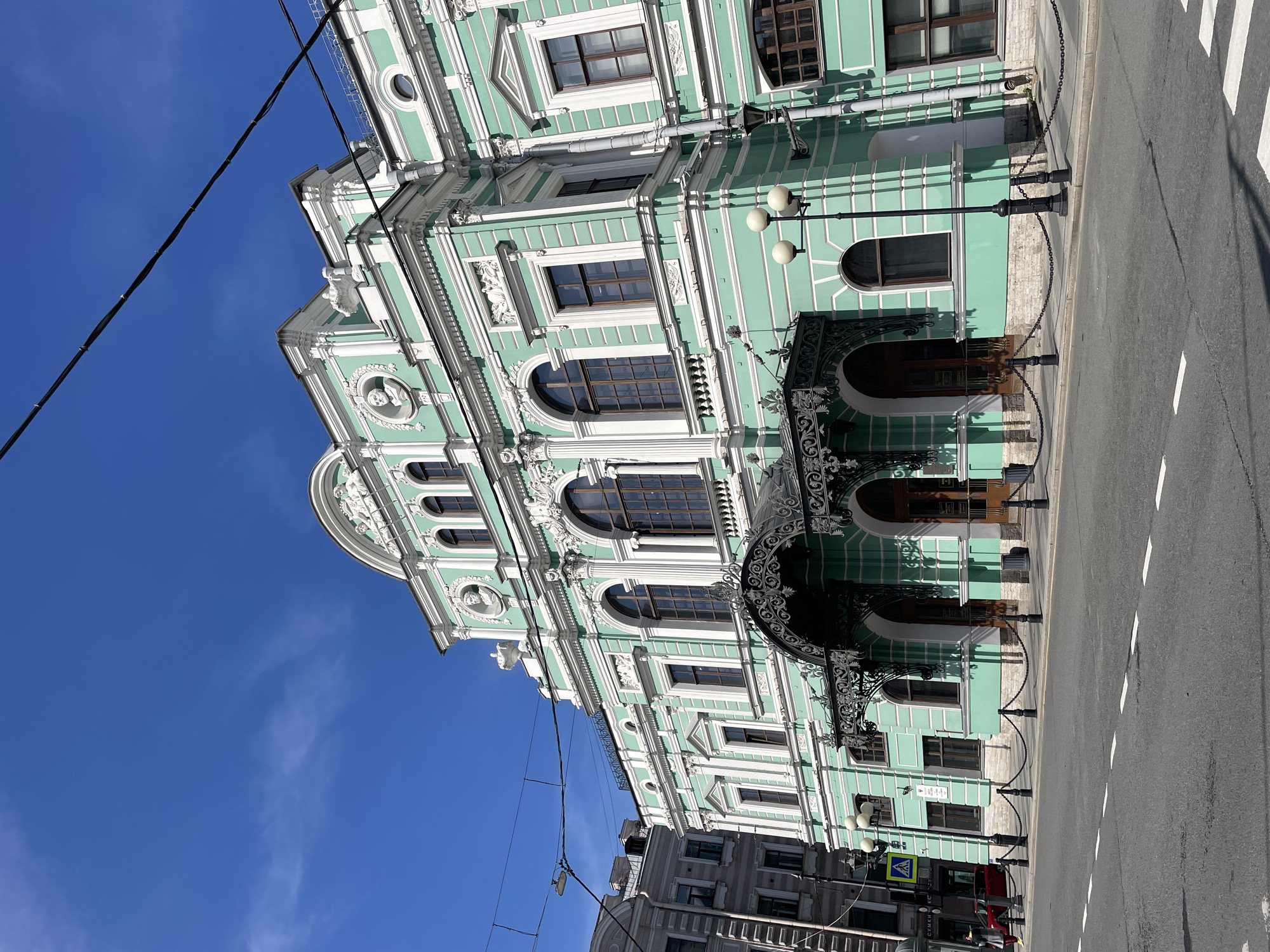 The image captures a daytime view of the Mikhailovsky Theatre in Saint Petersburg, Russia. The theatre's facade, painted in a pale green and white color scheme, dominates the frame. Its architectural details include decorative moldings, arched windows, and ornate balconies. In front of the theatre, a black wrought-iron canopy covers the main entrance. A chain-link fence runs parallel to the building along the sidewalk. A street with a zebra crossing and white lane markings stretches along the right side of the frame. Overhead, power lines crisscross against a clear blue sky. The lower portion of a neighboring building is visible in the bottom left, indicating an urban setting. People are visible on the street level near the crosswalk and other cars parked across the street, however their faces are indistinguishable.