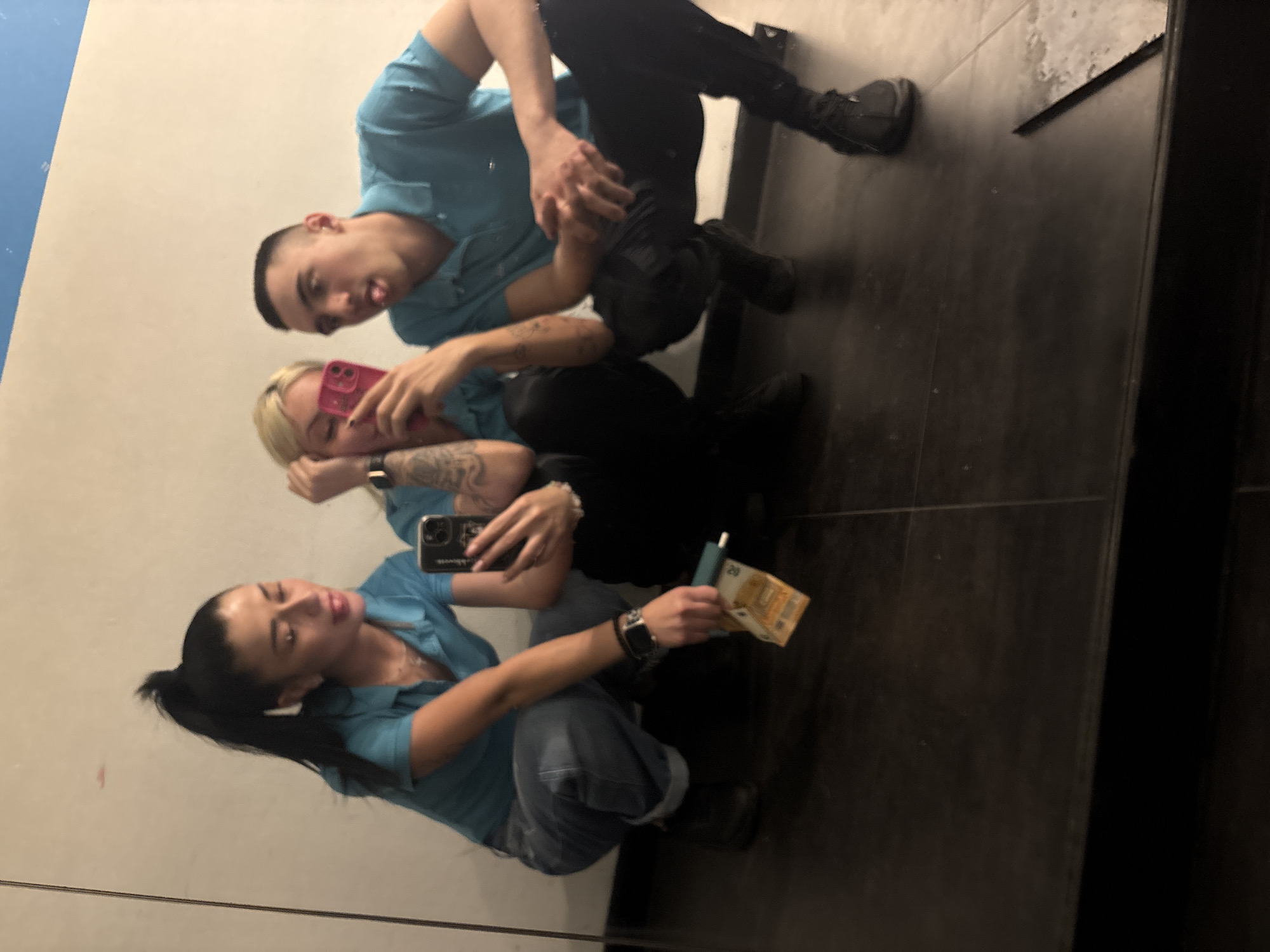 Three individuals are visible in a mirror's reflection. The person nearest the top of the frame is a male with short dark hair, wearing a light blue shirt and dark pants. He is sticking his tongue out. The second individual, a woman with blonde hair and tattoos, is holding a pink smartphone. She also wears a light blue shirt. The third person, a woman with black hair pulled into a ponytail, is holding a mobile phone and Euro bills. She is wearing a light blue shirt and jeans. All three people are squatting, presumably taking a selfie in front of a mirror that is located in a room with black tiles. The location is unknown, and there is no evidence it is in Plovdiv, Bulgaria.