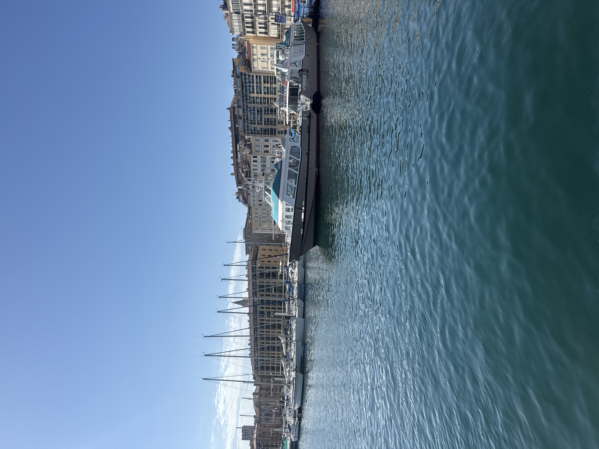 In a harbor, the still, deep blue water reflects the clear sky. Several sailboats with tall masts are docked in a line. Beyond the sailboats, two passenger ferries are moored. The ferry on the right is labeled "HYBRIDE." In the background, a row of multi-story buildings with arched windows lines the waterfront, suggesting an urban setting. The sky is a bright, cloudless blue.