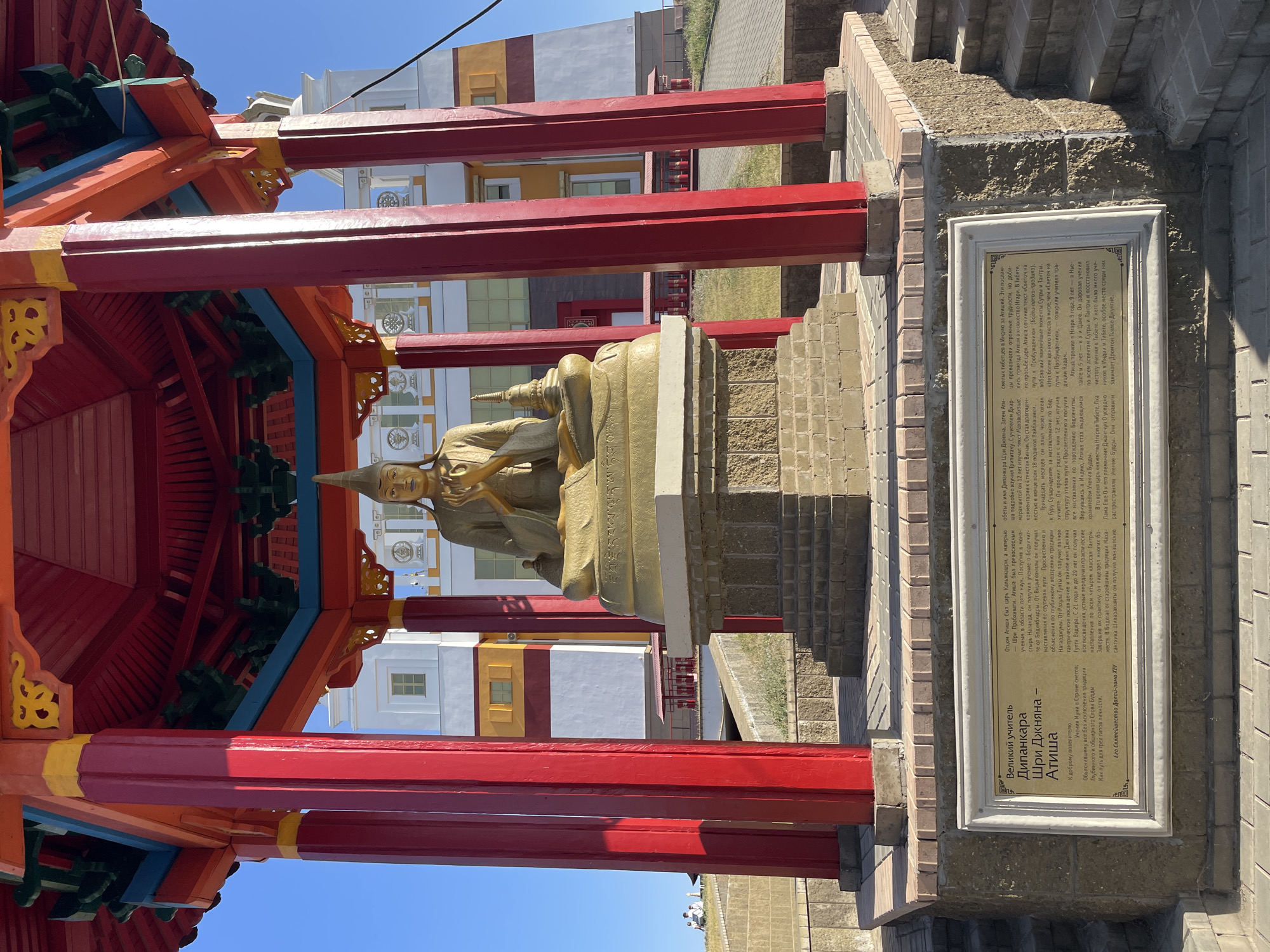 The image shows a golden statue of a seated figure, likely a Buddhist deity, under a red and yellow pagoda-like structure. The statue is positioned on a stone pedestal. In the background, a large white and yellow building with arched windows is visible. A plaque with text in Cyrillic script is mounted on a stone base in front of the statue. The environment appears to be outdoors, with a clear blue sky. There are a few people visible in the far background, sitting on a grassy slope. The overall scene is one of a religious or cultural monument. The text on the plaque, which appears to be in Russian, is associated with the city of Elista, Russia, and specifically mentions "The Great Teacher Djankara Shiri Djagna-Atisha," indicating a Buddhist context, consistent with Elista being the capital of the Republic of Kalmykia, known for its Buddhist population.