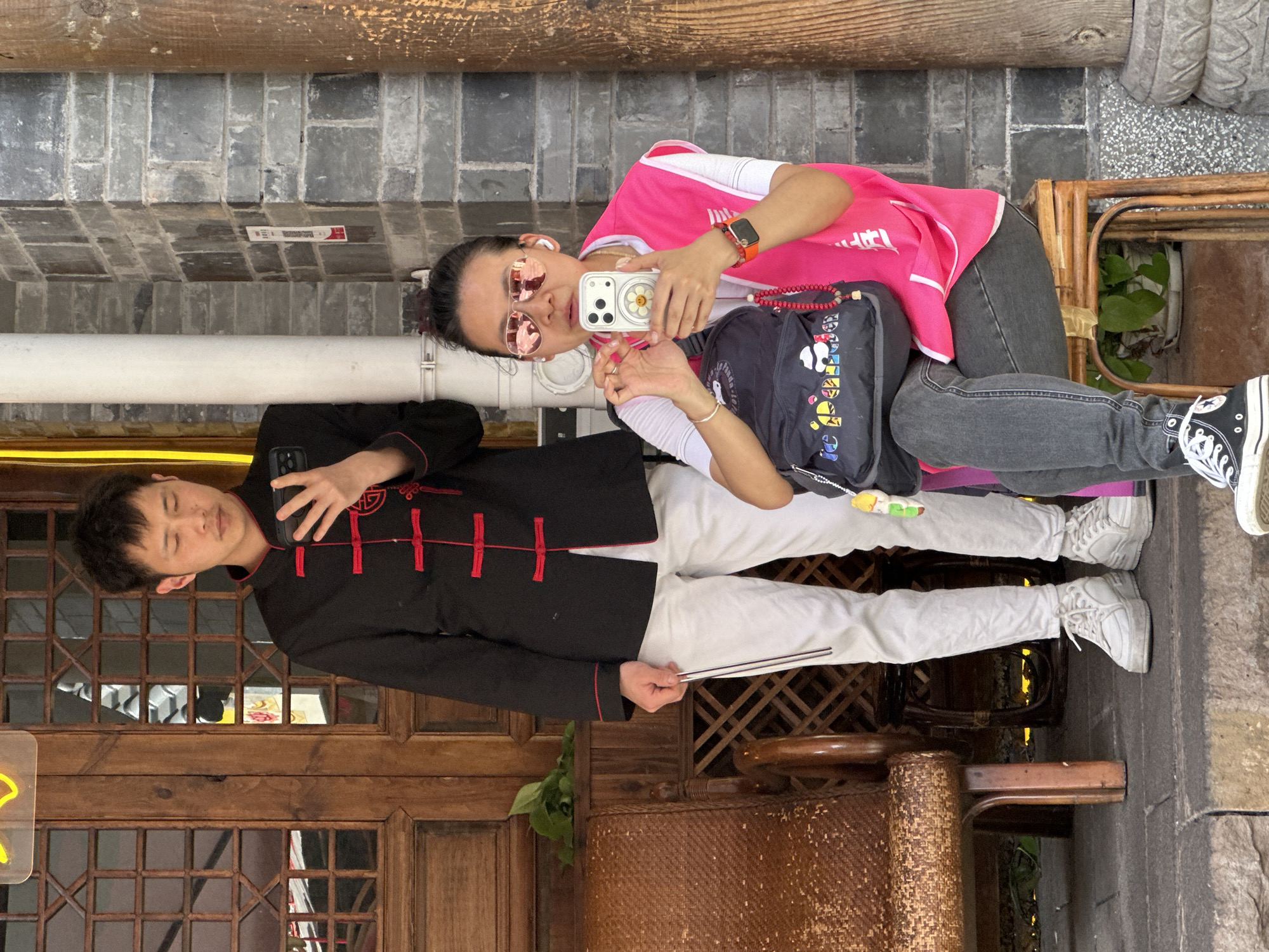 In Yuzhong District, China, a woman in a pink shirt and sunglasses is taking a picture with her smartphone of a man in a black traditional Chinese jacket with red trim. The man is holding a pair of chopsticks in his left hand and posing with his right hand over his heart. They are both seated in brown woven chairs on a stone walkway. Behind the man is a wooden door with latticework, and above them is a gray brick wall.