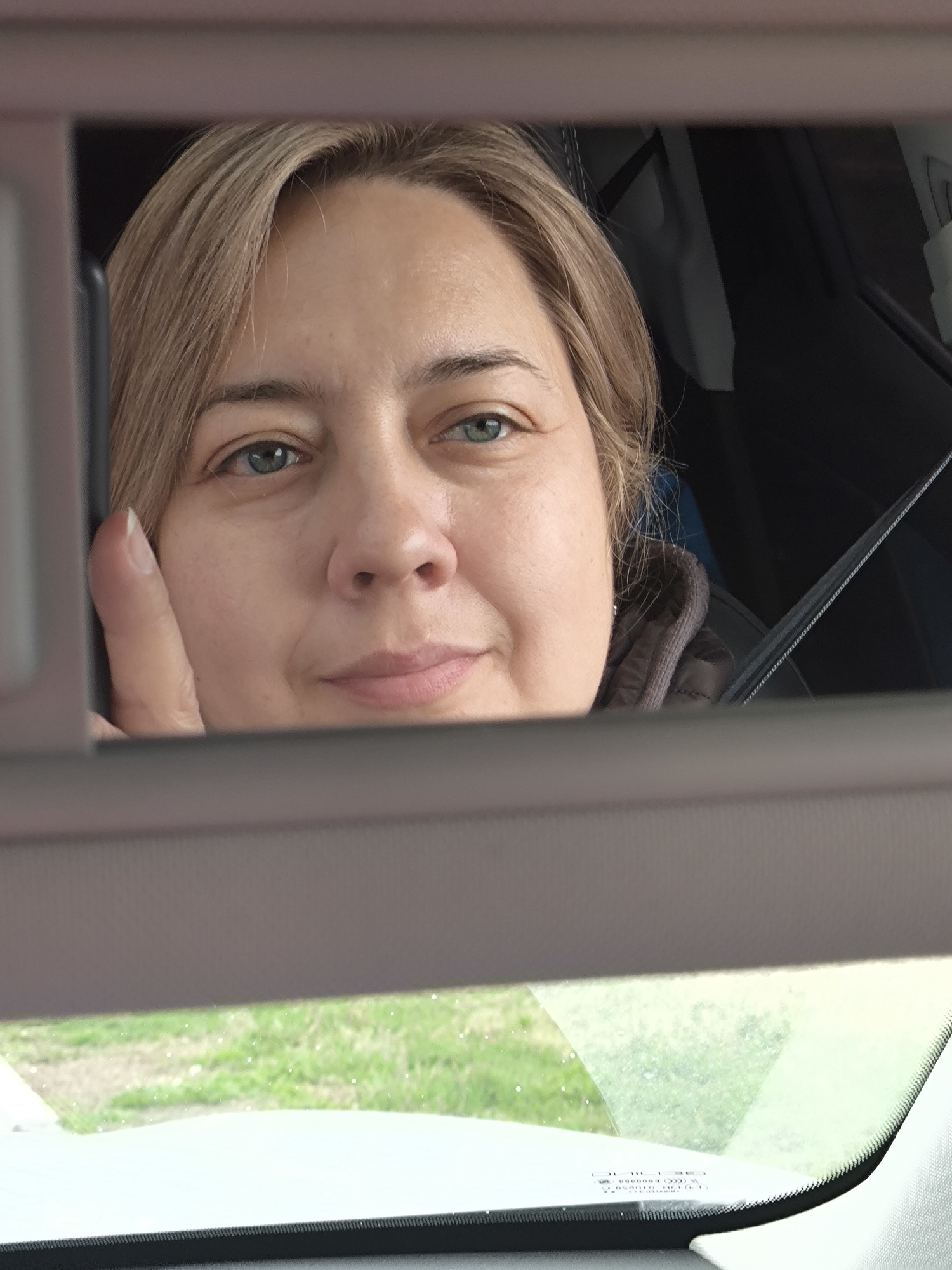 A woman with fair skin and light brown hair is reflected in a car visor mirror. The woman is looking slightly to her right with a subtle smile. Her thumb is visible on the left side of the mirror. Behind the visor mirror, a car interior is visible, including a dark seat and a seatbelt. Below the visor, the car windshield shows a view of green grass and what appears to be a road or sidewalk outside. There is no information to indicate the picture location is Belkino, Russia.