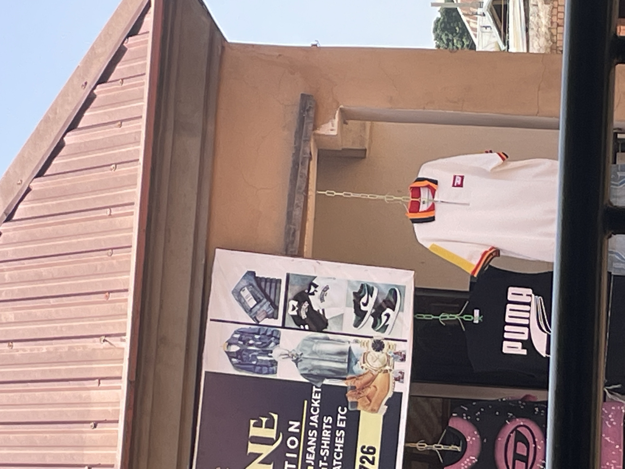In the city of Bukuru, Nigeria, a shop displays clothing and accessories. The shop, named "NE CTION", features a banner advertising various items like jeans jackets, t-shirts, and watches, along with phone numbers. The banner includes images of clothing, footwear, and timepieces, suggesting a retail establishment selling fashion items. In front of the shop, a white polo shirt with colorful accents hangs from a chain, and other clothing items like t-shirts and a jacket are visible. The setting appears to be an outdoor market or a street-side shop, with a corrugated iron roof overhead and a partly visible building in the background. The overall scene is a snapshot of a commercial area in Bukuru, Nigeria, showcasing local retail offerings.