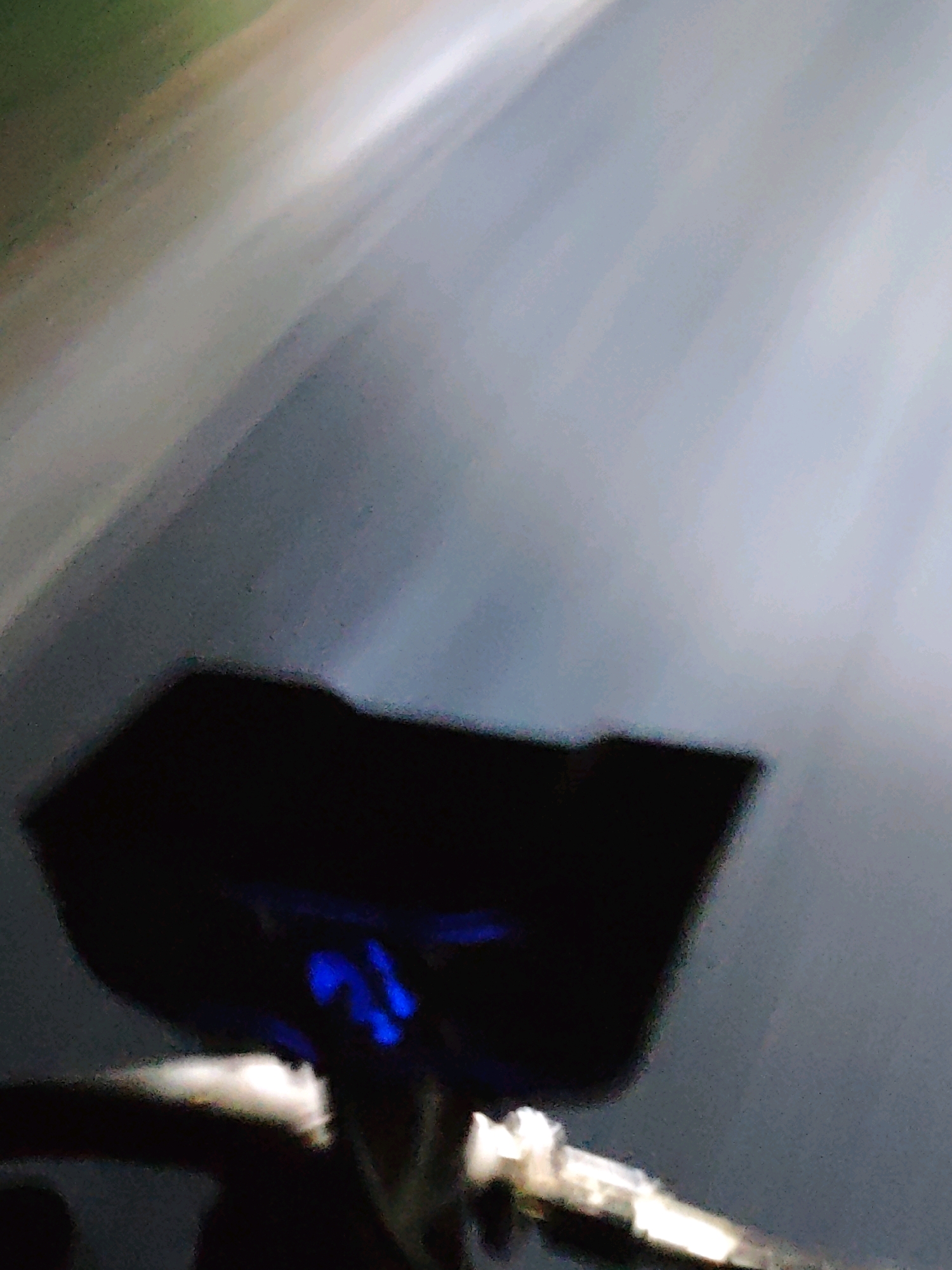 The image shows a blurred, dark object with some blue light emanating from it, positioned in the foreground. The background is also blurry, with streaks of light and muted colors suggesting a moving environment, possibly a road or pathway. The scene appears to be at night or in low light conditions. The location is identified as Muara Dua, Indonesia, though no specific landmarks or contextual details about the city are visible in the image itself.