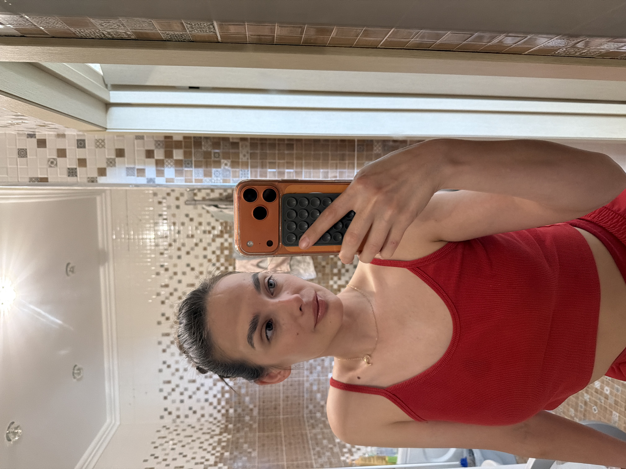 A young woman with dark hair pulled up in a bun and light-colored eyes takes a mirror selfie in a bathroom. She is wearing a red tank top and red bottoms. A gold necklace with a small charm is visible around her neck. Her right hand holds an orange-cased phone up to the mirror. The bathroom walls are tiled with a pattern of small brown and white squares, and there is a white ceiling with recessed lighting.