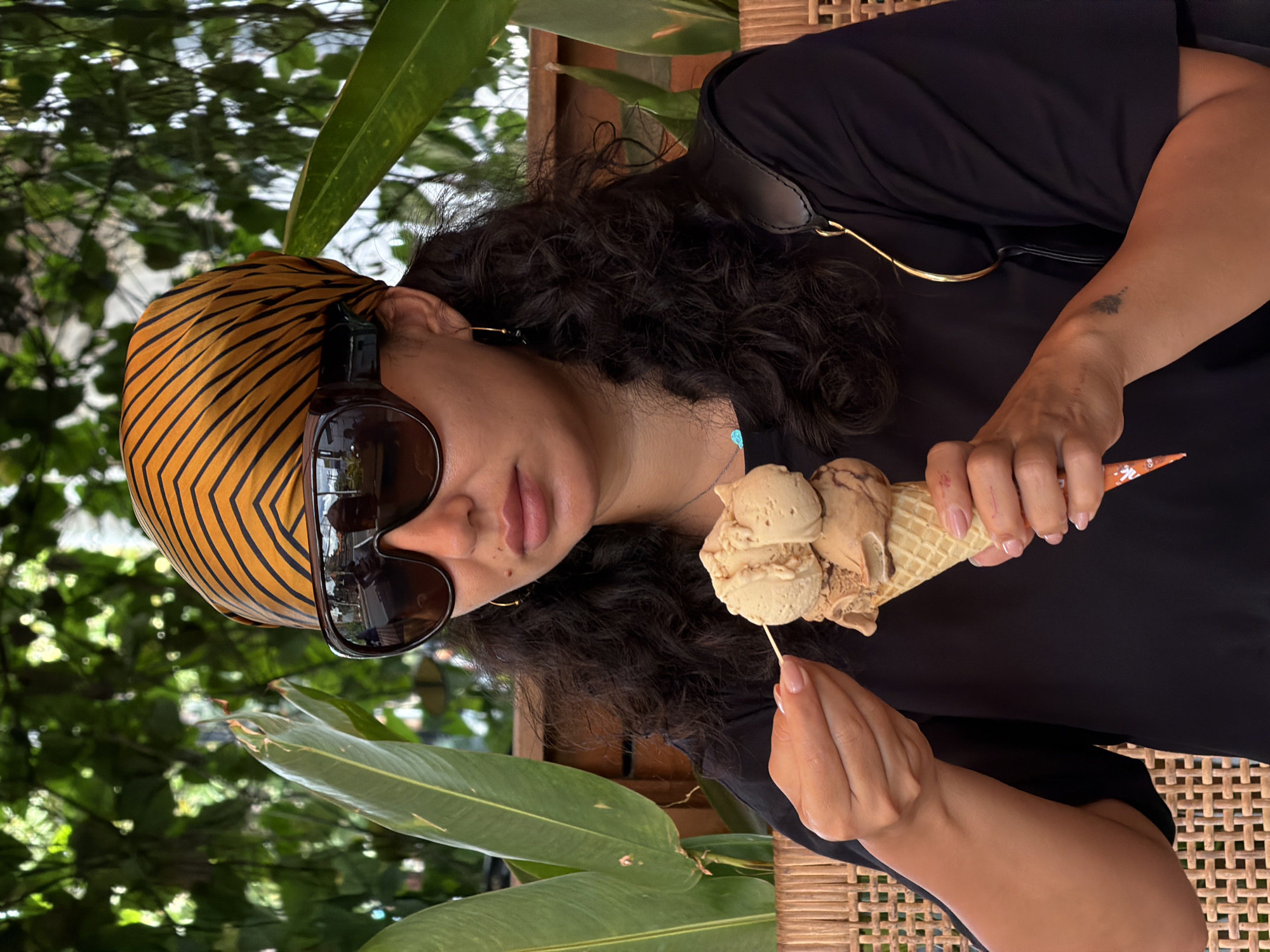 A woman is photographed in North Kuta, Indonesia, holding a cone with three scoops of light brown ice cream. She is wearing a headscarf with orange and black stripes and large sunglasses. She is sitting at a wicker table with tropical plants in the background. She is holding a small wooden stick, appearing to taste the ice cream. The woman is wearing a dark-colored, loose-fitting shirt.