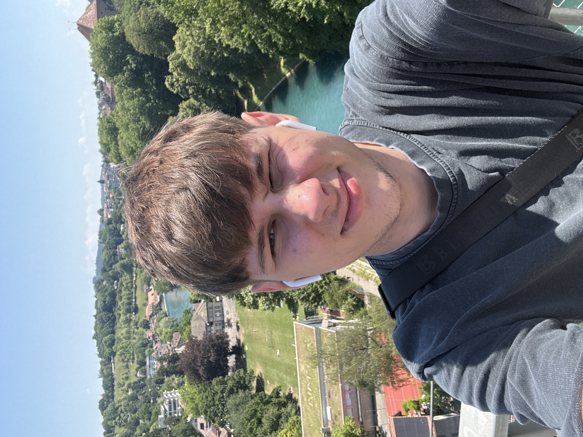 The image is a selfie of a young man with brown hair and light skin, wearing a dark grey t-shirt and white wireless earbuds. He is winking and smiling at the camera. He has a black strap from a bag crossing his chest.
The background shows a panoramic view of the city of Bern, Switzerland. A bright blue river flows through the city, with lush green trees and buildings lining its banks. In the distance, rolling hills are covered in dense foliage, under a clear blue sky with a few wispy clouds. The architecture of the buildings suggests a European city, and the specific landscape with the turquoise river is characteristic of Bern. The perspective suggests the man is standing on a bridge or an elevated viewpoint overlooking the city. The lighting indicates it is daytime, likely midday or early afternoon, with strong sunlight casting shadows.