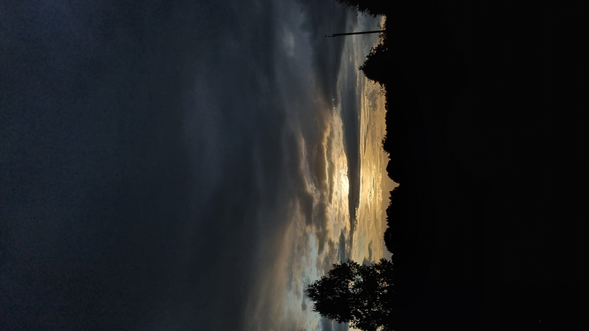 The image shows a dark, cloudy sky at dusk. The sun is setting and is visible through a gap in the clouds, casting a golden light. The foreground is silhouetted against the sky, with trees and a field visible. There is a telephone pole in the background on the right. The location is Vysokoe, Russia.