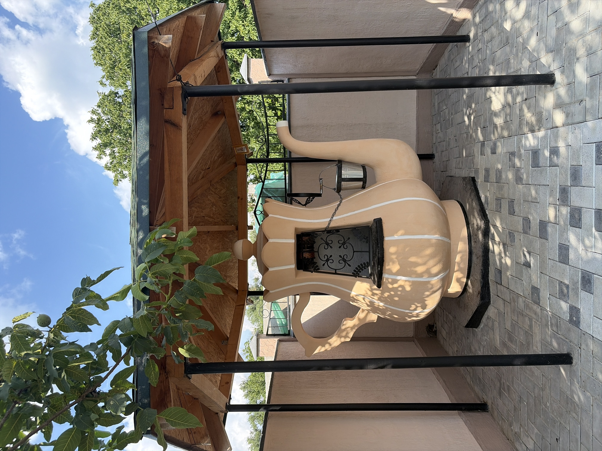 In Oxentea, Moldova, a large, beige, teapot-shaped oven is housed under a wooden gazebo with a green roof. The oven features decorative white stripes and a black wrought-iron window with an ornate scroll design. A metal bucket hangs from a chain on the side of the oven's spout. The structure is situated on a paved patio made of interlocking gray and black tiles. The background shows a light-colored wall, a metal fence, and lush green trees under a bright blue sky with scattered white clouds. Branches from a nearby tree hang over the gazebo.