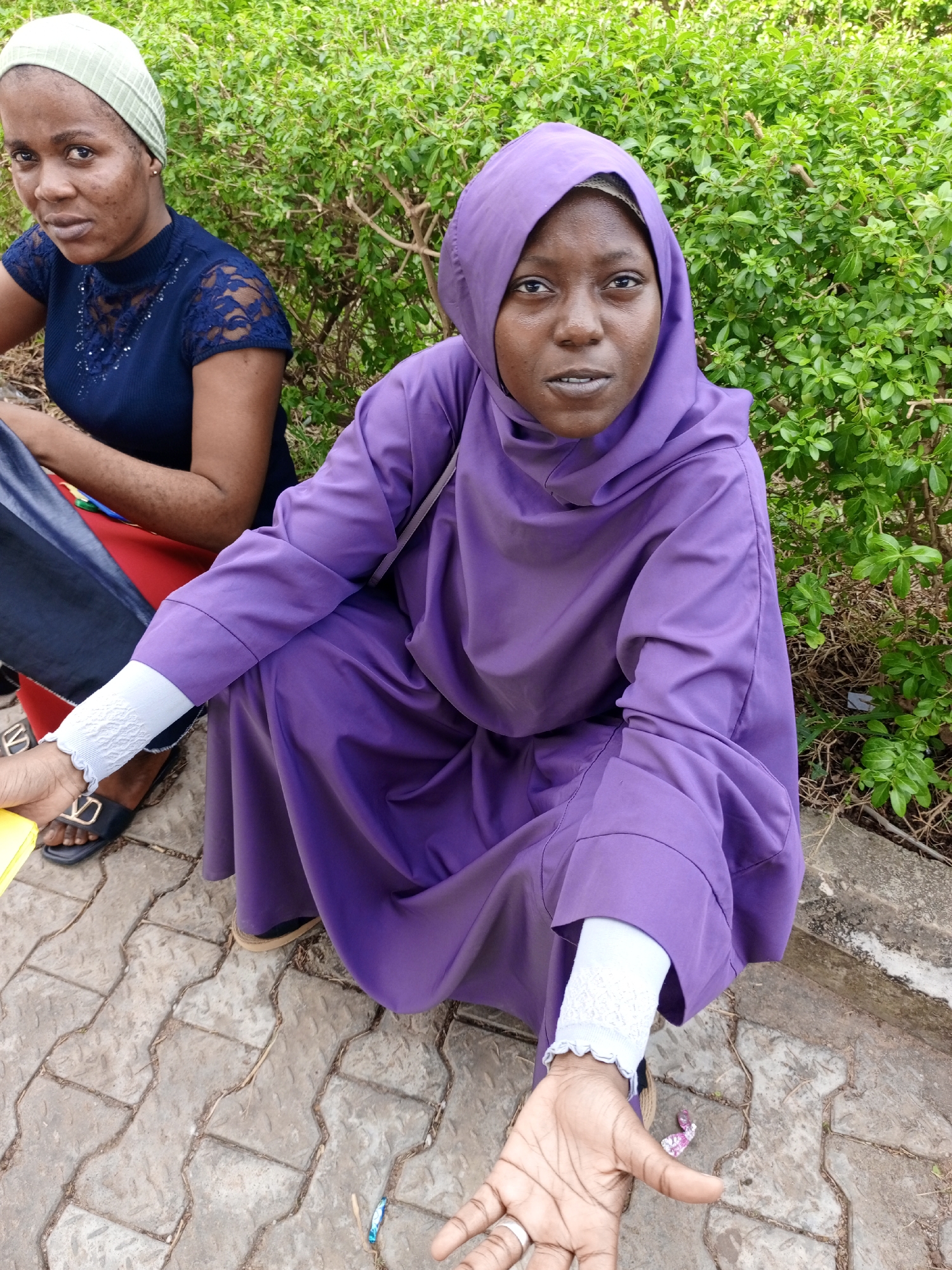 Two women are captured in an outdoor setting in Keffi, Nigeria. The focal point is a woman in a full-length, hooded purple robe. She is squatting, extending her open hand toward the camera, and wearing a ring on one of her fingers, with what appears to be a white under-sleeve with lace-like trim peeking out from under the robe. The ground is paved with patterned stones. To her left and in the background is another woman wearing a green head covering, a dark blue, short-sleeved top with lace detailing, and jeans. A section of a red garment is visible. Both women are dark-skinned. Lush green foliage fills the background.