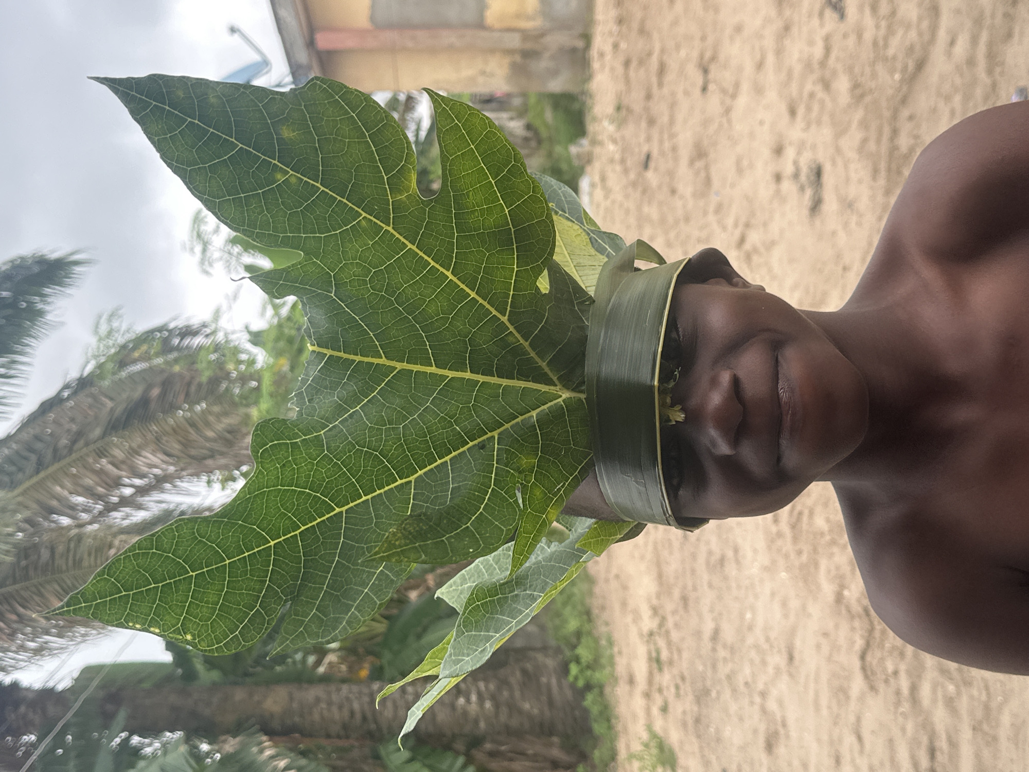 A young Black individual, likely a child, stands in a sandy outdoor area, wearing a headdress made of large green leaves and a band crafted from woven leaves around their forehead. The headdress prominently features a large, veined leaf that extends upwards, obscuring the top of their head. Smaller leaves sprout from the sides of the headdress. The individual has dark skin, short dark hair, and is looking directly at the camera with a slight smile. They are shirtless.
The background consists of sandy ground, a blurred building with faded colors and a satellite dish, and lush green vegetation including palm trees. The sky is overcast. The location is identified as Badagry, Nigeria.