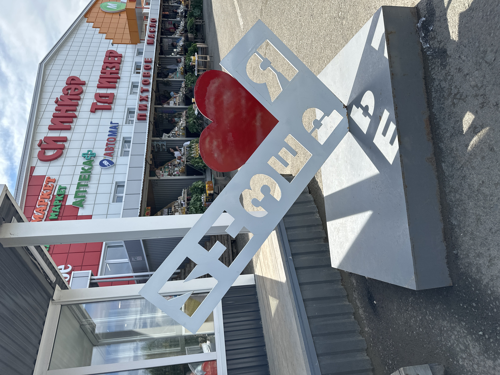 A frontal medium shot captures a large metal sculpture shaped like a diamond, with the Cyrillic letters "ИНЗЕР" cut out and a red heart placed behind the letters "ЗЕ". The sculpture is located in front of a building with the words "СЙ ИНЗЕР" prominently displayed in large red lettering. Below this, signs for "МАРКЕТ", "АПТЕКА+" and "АВТОМАТ" are visible, along with "ПИХТОВОЕ МАСЛО". Stalls are set up under a canopy in front of the building, displaying goods, and people can be seen browsing. The sculpture is situated on a paved surface, with shadows cast by the sun. The overall scene appears to be an outdoor market area in the city of Inzer, Russia.