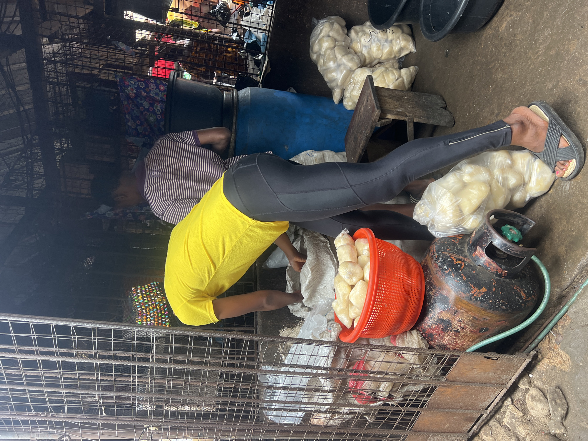 In Nkpor, Nigeria, a person wearing a bright yellow t-shirt and black leggings is bent over, reaching into a white sack. Beside them, a red basket overflows with pale, starchy items. A large, rusted gas cylinder lies horizontally in the foreground, connected to a green hose. Behind the person in yellow, another individual in a striped shirt stands near a large blue barrel and a dark tub. Bags of similar starchy items are stacked in the background. The scene appears to be a market stall, with metal bars and a rough, dirt floor suggesting an outdoor or semi-enclosed environment.