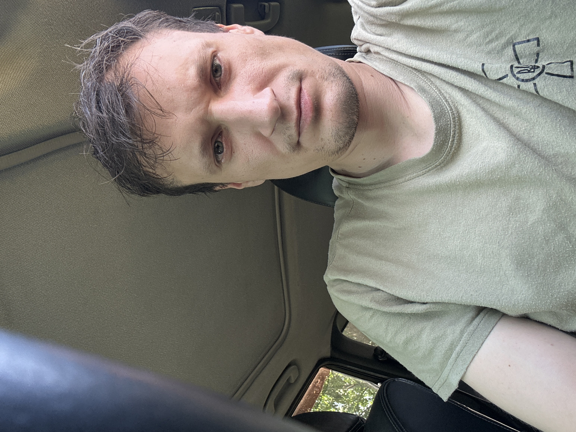 The image is a selfie taken from inside a vehicle. The subject is a man with light skin, brown hair, and a neutral expression. He is wearing a light-colored t-shirt with a black emblem on the left side of his chest. The interior of the car is visible, including the sunroof, the dashboard, and part of a black car seat. Through the window, trees and foliage can be seen, suggesting an outdoor location. The image does not contain any information that would place the location specifically within the city of Makiv, Ukraine.