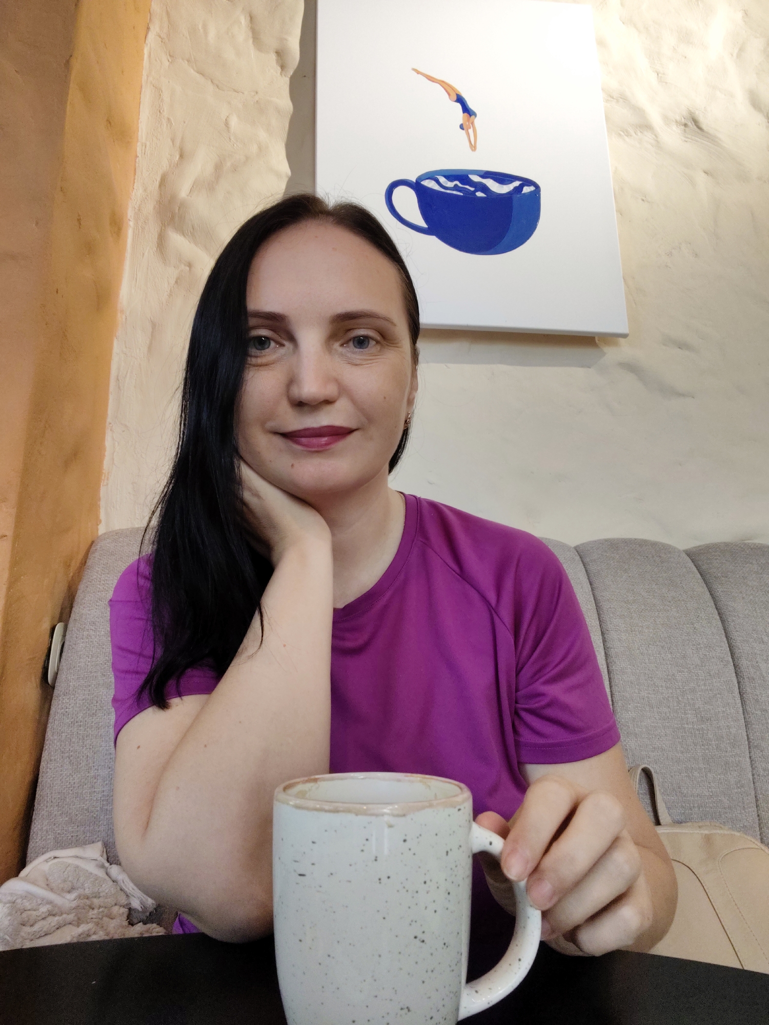 A woman with long dark hair is seated on a grey tufted sofa in what appears to be a cafe, possibly located in Sochi, Russia. She is wearing a vibrant purple t-shirt and has her left arm resting on her chin. In her right hand, she is holding a speckled white ceramic mug. Behind her, on a white wall, is a piece of art depicting a person diving into a blue coffee cup. The cafe has textured beige walls and a dark table in the foreground.