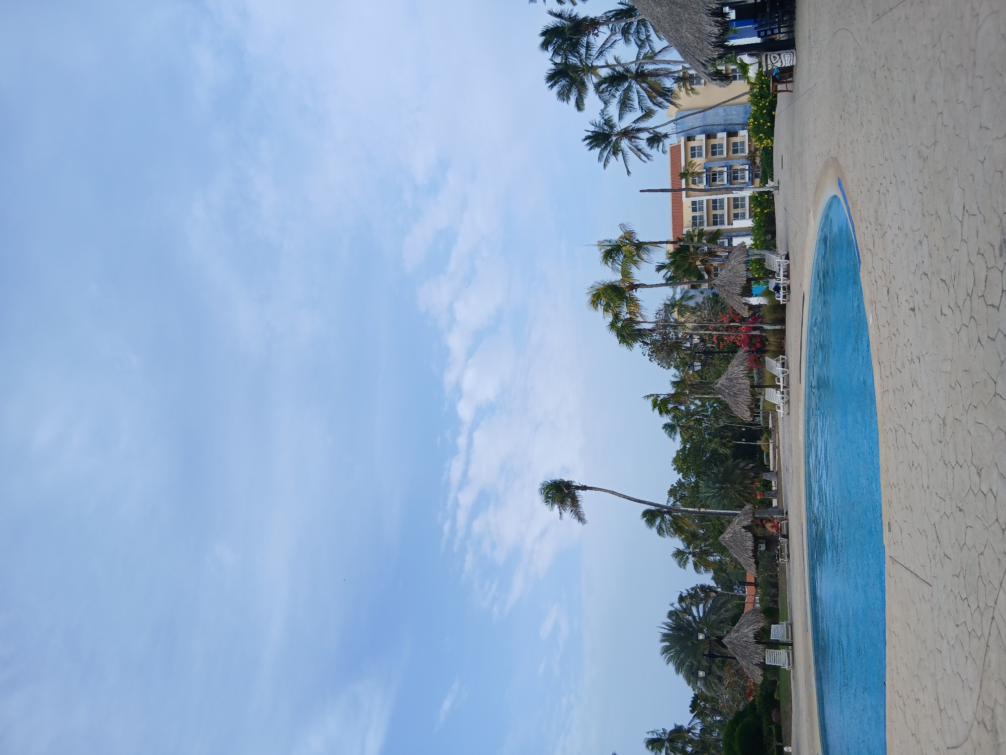The image captures a scene in Playa El Agua, Venezuela. It features a view that includes a swimming pool with blue water, a beige-colored concrete patio surrounding the pool, and a backdrop of palm trees with thatched umbrellas. A multi-story building with blue and white exterior can be seen in the background. The sky above is light blue with scattered clouds. Several white lounge chairs are also visible, suggesting a resort or leisure setting.