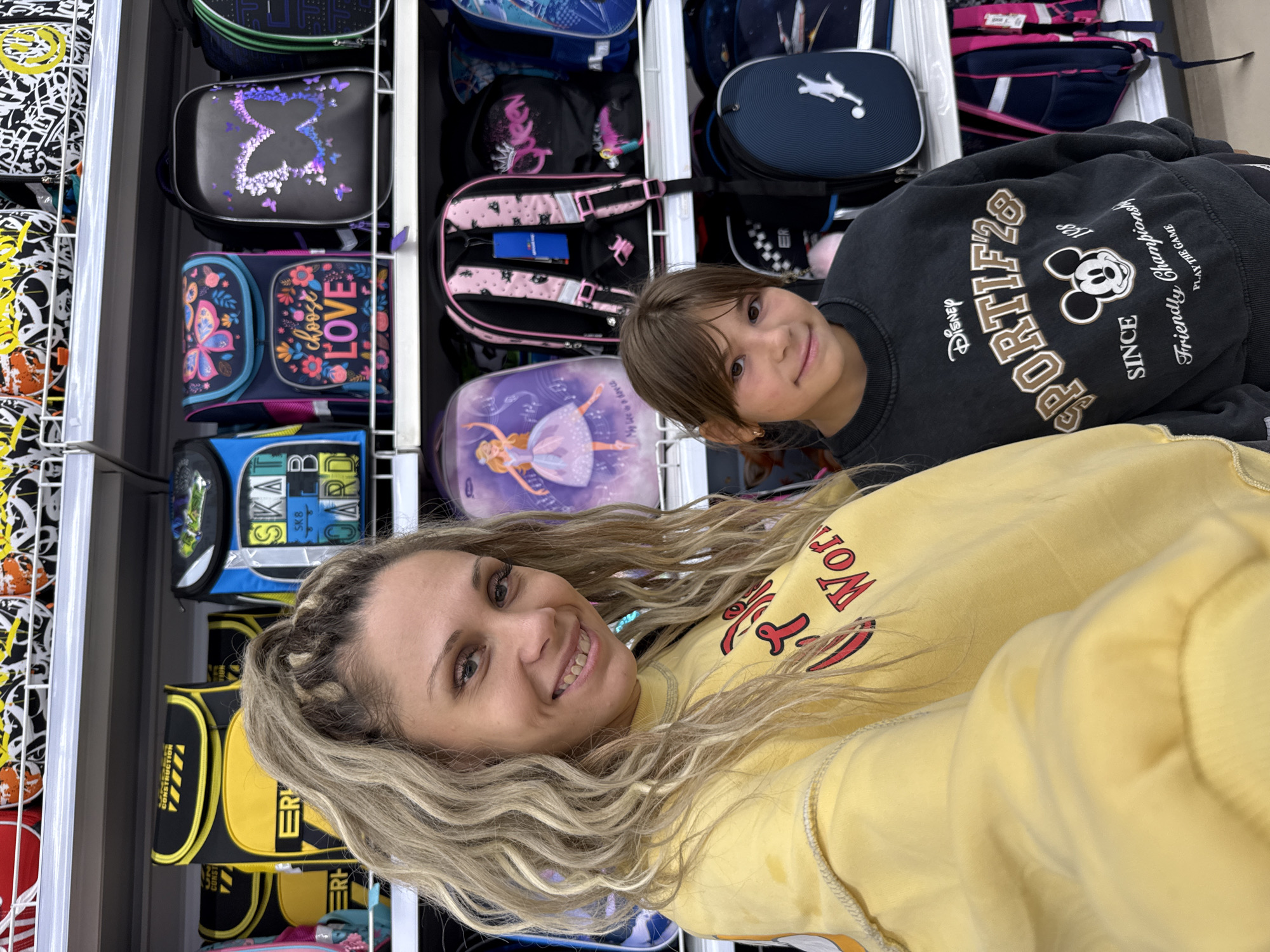 In Dumino, Russia, a woman with blonde, wavy hair and a yellow sweatshirt takes a selfie with a young girl wearing a dark grey sweatshirt. The woman is smiling at the camera, while the girl looks directly at it. They are standing in front of shelves filled with colorful backpacks, many of which are decorated with various designs and characters, including butterflies, ballerinas, and a Mickey Mouse graphic on the girl's sweatshirt. The overall environment appears to be a retail store.