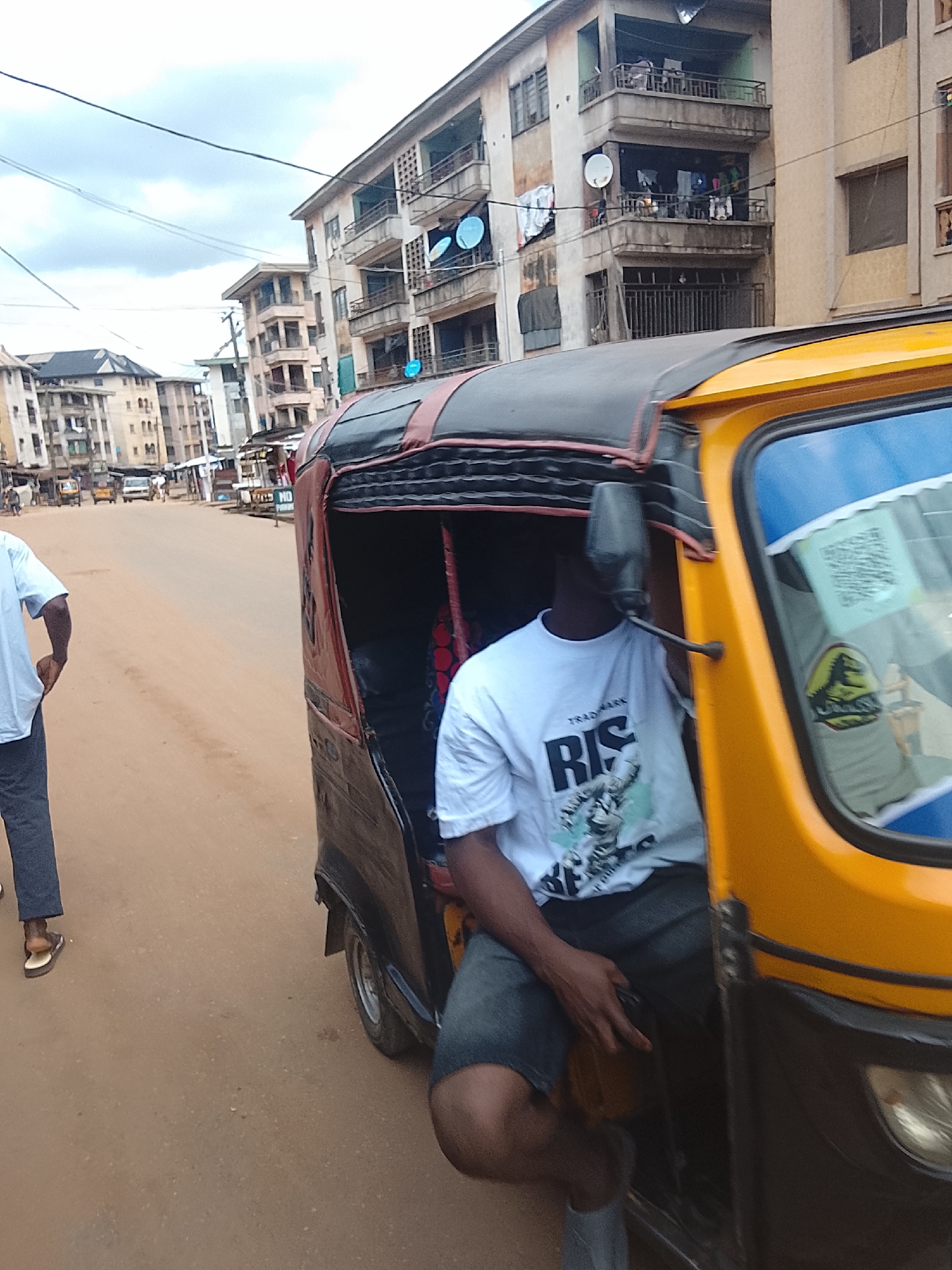 The image shows a street scene, likely in a developing country, with a yellow and black auto-rickshaw in the foreground. A person wearing a white t-shirt with the text "RIS" and graphics, and dark shorts, is seated in the auto-rickshaw. Another person, wearing a light blue shirt and dark pants, stands to the left of the auto-rickshaw, facing away from the camera.
Behind the auto-rickshaw, several multi-story buildings are visible, suggesting an urban or semi-urban environment. Some buildings appear older and in disrepair, with visible laundry hanging from balconies. The street is unpaved and dusty. In the background, other vehicles, including what appear to be cars and more auto-rickshaws, can be seen on the street. A sign with "NO PARKING" is visible on the right side of the street.
The sky is overcast with clouds, suggesting it might be a cloudy day. The lighting indicates it is daytime, but the exact time is difficult to determine. The overall impression is of a busy street in a lively but not necessarily affluent area. The presence of the auto-rickshaw and the style of the buildings are strong indicators of the location's context, likely in a region where such vehicles are common modes of transportation.