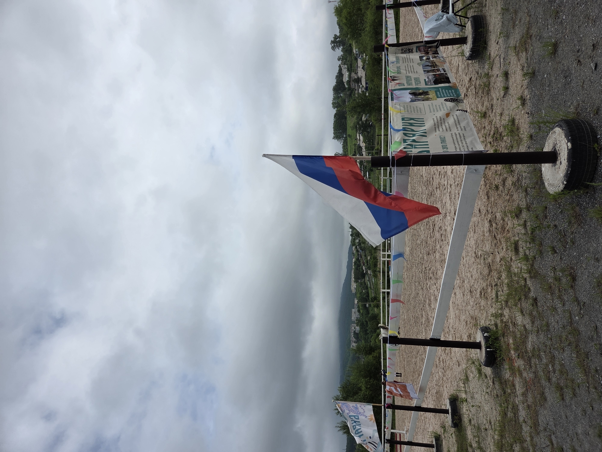 The image depicts an outdoor equestrian arena with a sandy ground, likely located in the city of Теплоозёрск, Russia. A Russian flag is prominently displayed, fluttering in the wind, positioned to the right of the frame. In the background, a chain-link fence encloses the arena, decorated with colorful triangular pennant flags. Beyond the fence, a gentle slope is visible, covered with green trees and dotted with buildings, suggesting a semi-rural or suburban setting. The sky above is overcast with heavy, dark clouds, hinting at potential rain or stormy weather. Two flagpoles stand within the arena, one supporting the Russian flag and the other holding a large banner with text and images, appearing to be informational or promotional in nature. Some smaller flags and banners are also visible along the fence line. The ground in the foreground is covered with gravel and sparse green grass. No individuals are clearly visible or engaged in any activity within the arena, though a horse might be present in the distance. The overall impression is that of a somewhat somber, cloudy day at an outdoor venue, possibly awaiting an event.