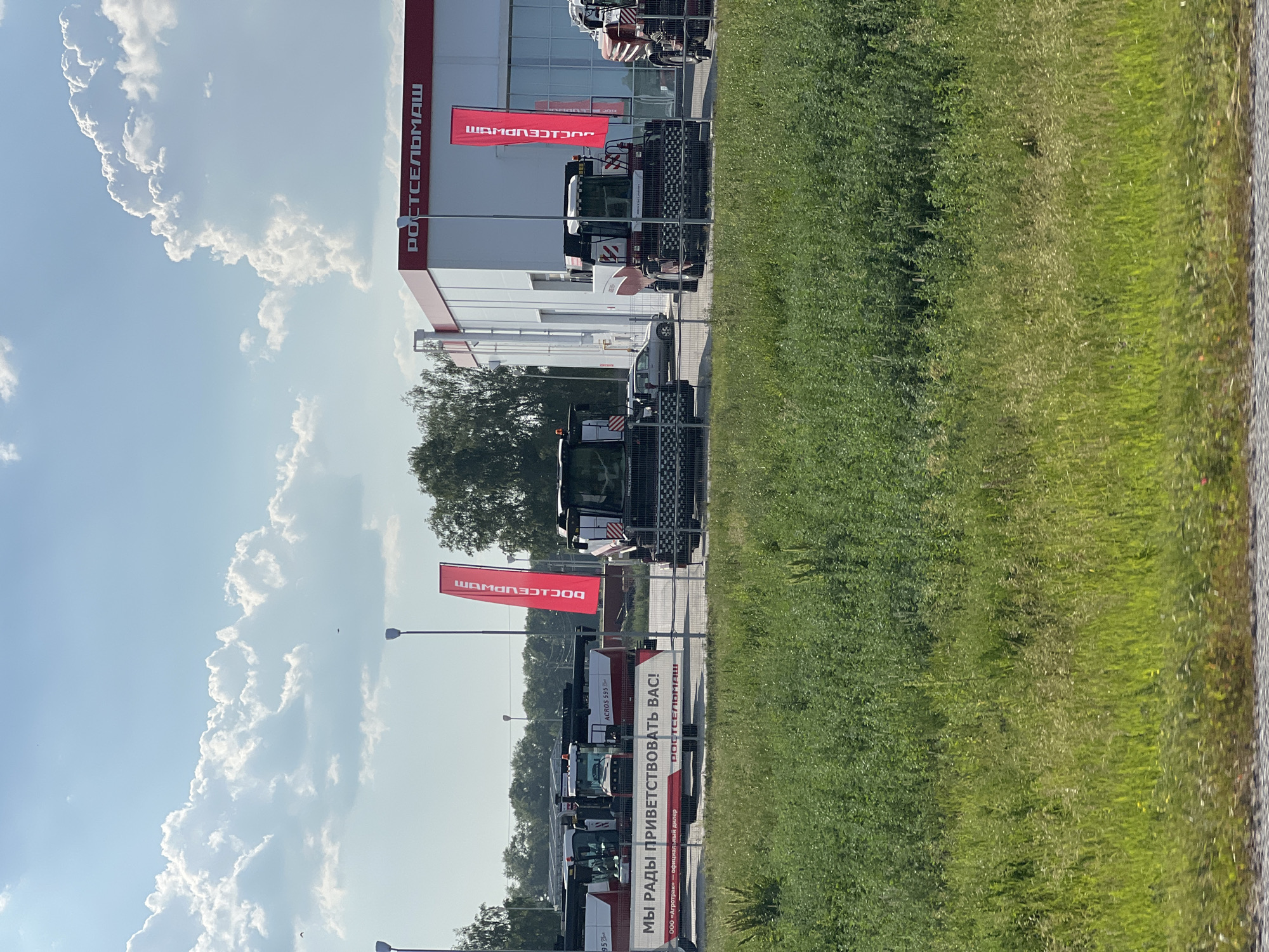 The image depicts an outdoor agricultural machinery dealership on a sunny day with scattered clouds. The foreground is dominated by a lush green grassy field. In the midground, behind a metal fence, several large agricultural machines, specifically harvesters and tractors, are parked. These machines are primarily white and red, with some bearing logos. A prominent red banner with white text in Cyrillic, reading "РОСТСЕЛЬМАШ," is visible on the right side of the image. Adjacent to the machinery is a modern building with a red-trimmed roof, also featuring the "РОСТСЕЛЬМАШ" logo in white letters. A smaller red banner with similar Cyrillic text is attached to a lamppost in front of the building. A car is parked to the left of one of the harvesters. The background consists of trees and a bright blue sky with large, fluffy white clouds. The text on the banner in front of the machines reads "МЫ РАДЫ ПРИВЕТСТВОВАТЬ ВАС!", meaning "WE ARE GLAD TO WELCOME YOU!". The text on the smaller banner reads "РОСТСЕЛЬМАШ" vertically. The scene suggests a commercial display of agricultural equipment. Based on the company name "РОСТСЕЛЬМАШ" and the nature of the equipment, this is likely a dealership located in Russia.