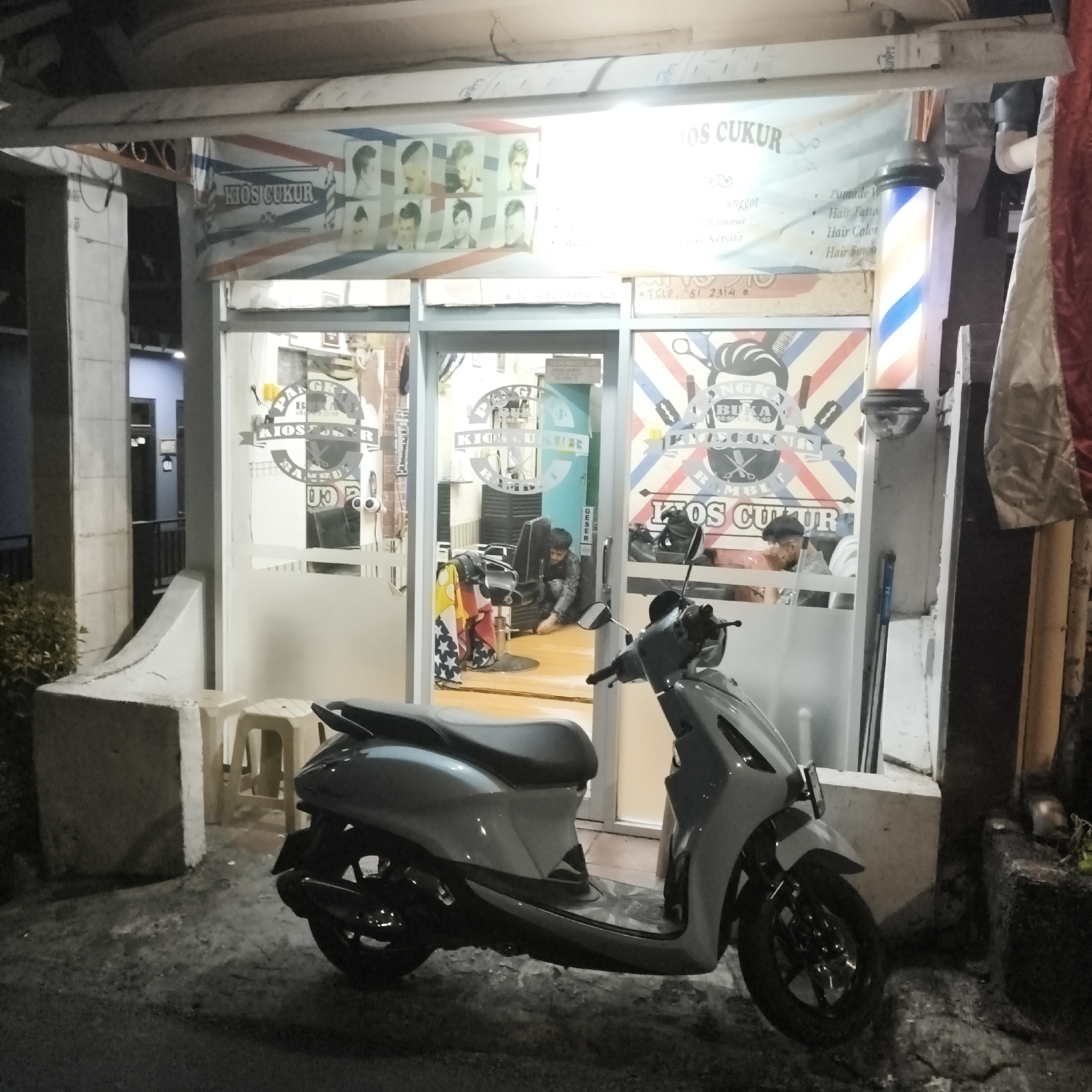 The image depicts the exterior of "KIOS CUKUR", a barber shop in South Jakarta, Indonesia, at night. A light blue and white scooter is parked in front of the shop. The shop features a large barber pole with red, white, and blue stripes, illuminated by light. A banner above the entrance displays various men's hairstyles and the text "KIOS CUKUR". The shop's glass door reveals a barber chair and a barber attending to a customer inside. Another customer is seated in the background. The street in front of the shop is paved and dimly lit.