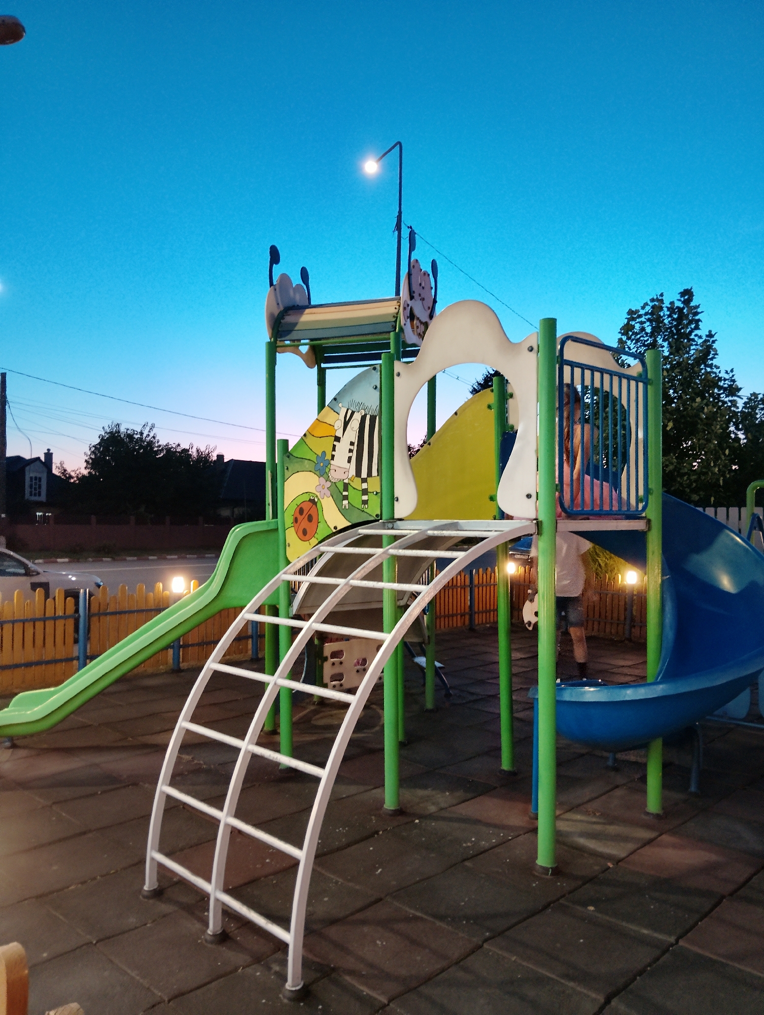 The image shows a children's playground in Adjud, Romania, at dusk. The playground features a large, multi-colored climbing structure with green slides, a blue slide, and a white arched climbing ladder. The structure is adorned with cartoonish illustrations, including a zebra and a ladybug. A small child, partially visible and wearing a pink shirt and shorts, is standing within the structure. The playground has a rubberized surface for safety. In the background, a wooden fence separates the playground from a street where a car is passing. Residential buildings and trees are visible behind the fence. The sky is a gradient of deep blue to pale pink, indicating twilight. Streetlights are illuminated, casting a warm glow on the scene.