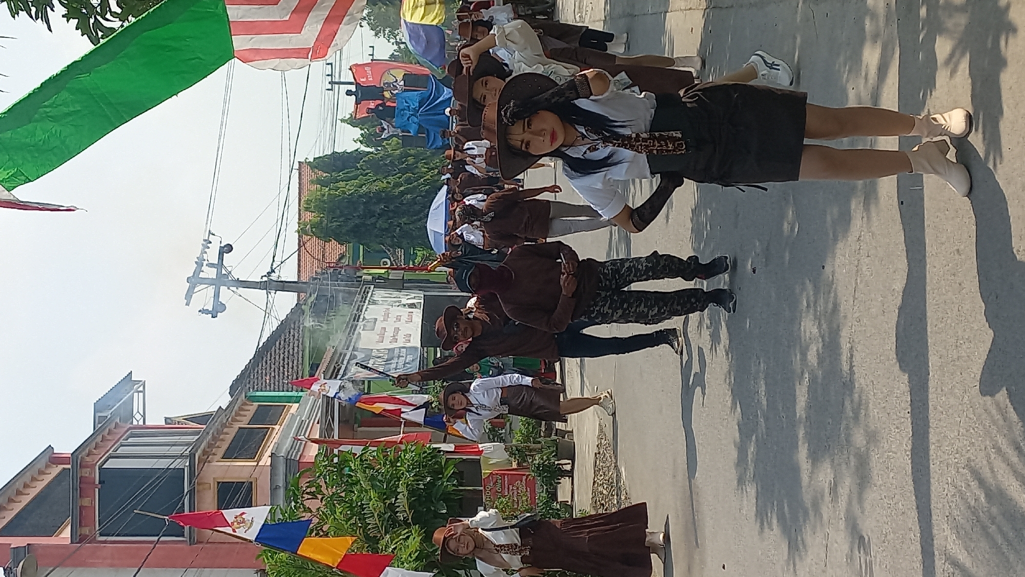 In the city of Kedungmegarih, Indonesia, a procession moves down a paved street under a clear sky. The procession features a group of people dressed in cowboy-inspired attire, including hats and brown clothing. Some individuals are holding flags and what appear to be props, such as a gun-like object. In the background, buildings with colorful facades and a utility pole are visible. Trees and foliage line the street. Several flags, including red and white ones, are present. The ground is covered in sunlight and shadows from trees, indicating an outdoor daytime event.