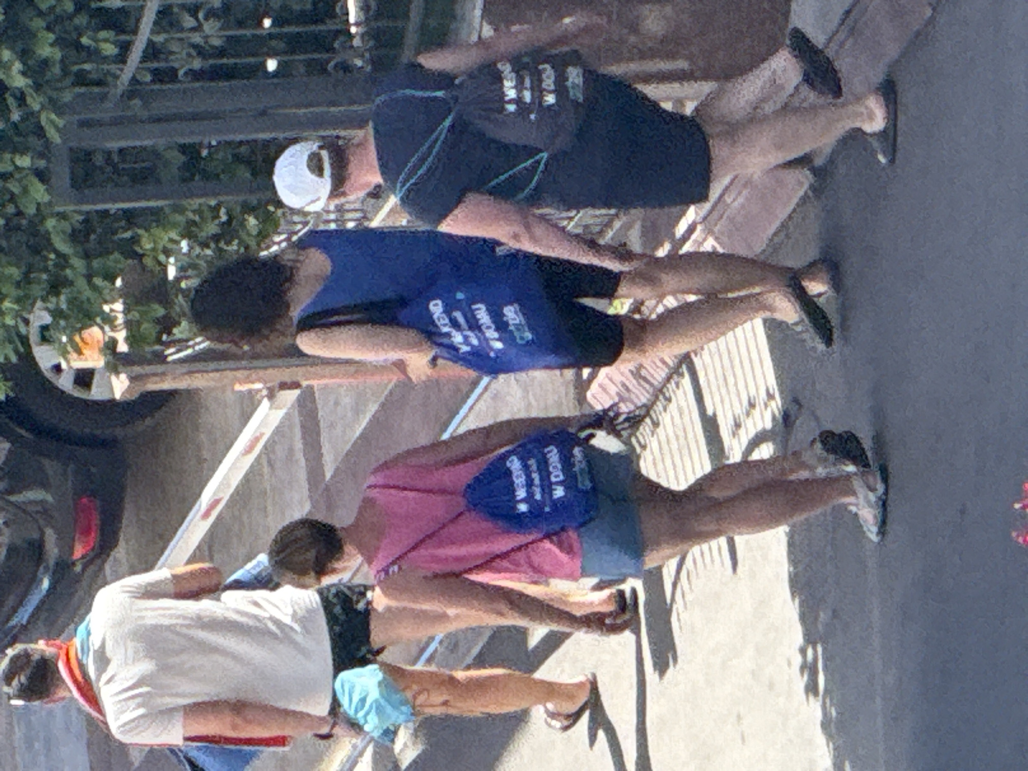Four adults walk away from the camera on a sunlit paved pathway in Budva, Montenegro. From left to right: A male with dark hair wears a white t-shirt, dark patterned shorts, and flip-flops, carrying a multi-colored towel over his left shoulder and a light blue item in his hand. Next, a female with short dark hair wears a pink tank top, light denim shorts, and athletic shoes, carrying a blue drawstring backpack with the visible Polish text "WEEKEND W DOMU Grabie." Following her, a female with dark curly hair wears a dark blue sleeveless top, dark cycling shorts, and dark flat shoes, carrying a blue tote bag with the same Polish text "WEEKEND W DOMU Grabie." Finally, an individual, likely female, wearing a white baseball cap backward, a dark short-sleeve top, and dark shoes, carries a dark gray drawstring backpack also displaying the Polish text "WEEKEND W DOMU Grabie." In the background, a white and red striped barrier is visible to the left, with part of a dark vehicle and its red taillight behind it. To the right, a dark metal fence with decorative elements is partially obscured by green foliage under bright sunlight, casting distinct shadows on the ground.