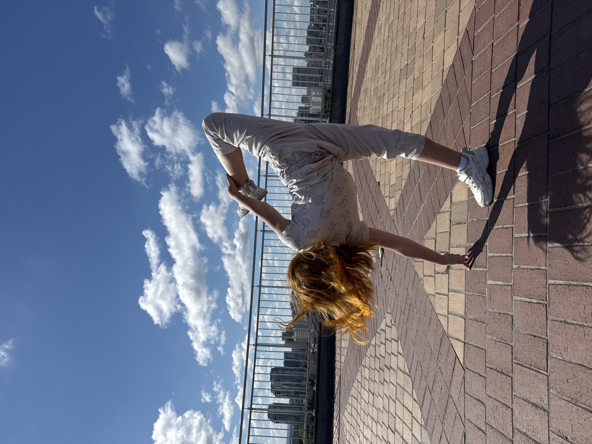 The image shows a person performing an acrobatic or yoga pose, specifically a backbend or bridge, in an outdoor urban setting. The subject, with long, flowing reddish-blonde hair, is upside down with their head hanging towards the ground. They are dressed in light-colored, loose-fitting pants, a patterned top with small dark designs, and white sneakers with white socks. Their hands are positioned near their feet, suggesting the execution of the pose.
The setting is a paved walkway or promenade with a distinct pattern of rectangular bricks in shades of brown and beige. A metal railing runs along one side, separating the foreground from a panoramic view of a city skyline. Numerous modern buildings are visible in the distance, silhouetted against the bright sky. The sky is a vivid blue, adorned with scattered white, fluffy cumulus clouds, indicating clear and sunny weather. Strong, elongated shadows are cast on the paved ground, suggesting a high sun angle, likely late morning or early afternoon. The location is Kazan, Russia. No visible text is present in the image.