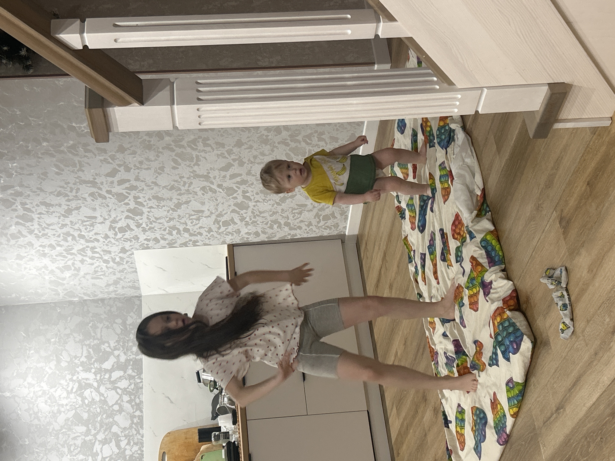 The image, taken in Chaikovsky, Russia, captures a casual indoor scene featuring a woman and a young child. The room appears to be a modern home interior with light wood laminate flooring and light-colored walls, one of which has a textured or patterned wallpaper in shades of grey and white.
In the foreground, a woman with long dark hair is standing, seemingly mid-action, possibly dancing or posing playfully. She is wearing a light-colored, short-sleeved top with a subtle pattern and grey athletic shorts. Her arms are slightly raised, and her body is angled. Behind her, light-colored kitchen cabinets and a countertop are visible, holding items like a wooden cutting board and what appears to be a smartphone in a green case.
To the right, a young child, a toddler with light hair, stands beside a white, multi-tiered wooden play structure or furniture piece. The child is dressed in a yellow t-shirt featuring a banana graphic and green shorts or a diaper cover. They are looking towards the camera with a neutral expression. On the floor beneath the child is a large, colorful blanket or mat adorned with numerous "Pop It" fidget toy designs. A single crumpled grey sock with patterns lies on the floor next to the blanket.
The lighting suggests it is daytime, providing a bright and clear view of the scene. No discernible text is visible within the image.