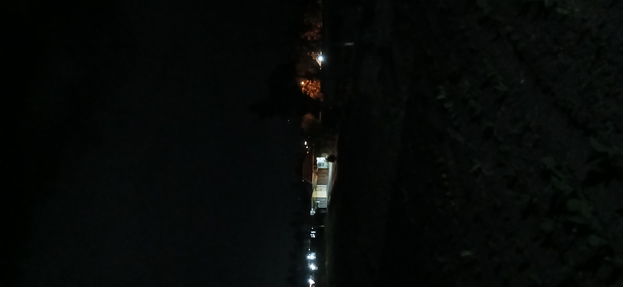 The image depicts a nighttime scene in Sekarputih, Indonesia, showing a dimly lit rural area. In the background, a single-story building with a tiled roof is visible, illuminated by interior lights. A person is walking away from the building on a pathway. Distant lights suggest the presence of other buildings or streetlights in the vicinity. The foreground is dark and appears to be an open field or uncultivated land. Trees are scattered throughout the scene, with some illuminated by lights, casting faint shadows. The overall atmosphere is dark and quiet, characteristic of a rural setting at night.