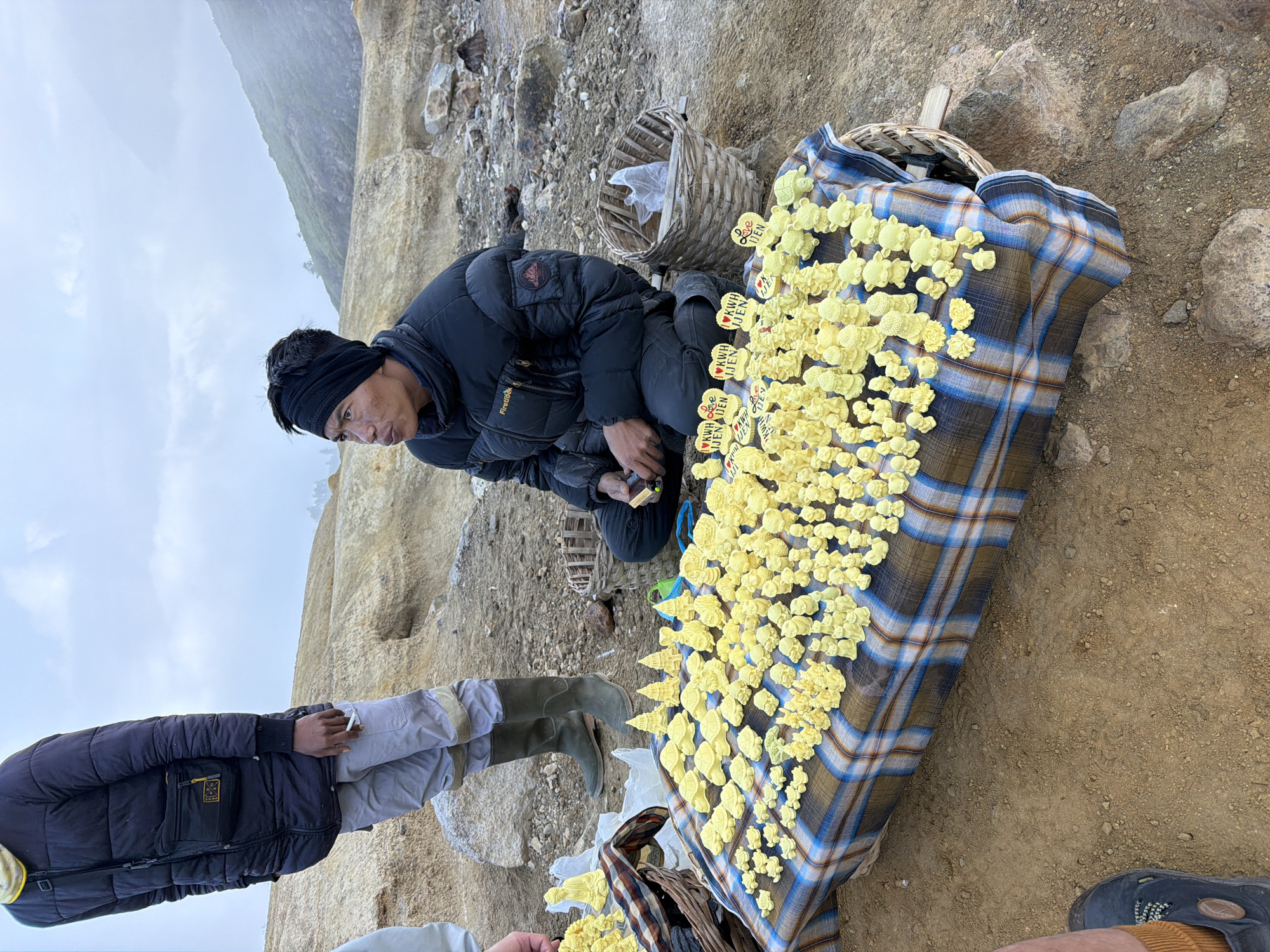 A vendor kneels on rocky ground in a misty, mountainous environment, displaying numerous small, yellow, sulfur-carved figures on a checkered cloth spread over a makeshift table. The figures are diverse, depicting animals like turtles and bears, as well as Buddha statues and trees. Some figures have heart-shaped tags attached, reading "Ijen" and "Love Ijen." The vendor wears a black puffer jacket, a black headwrap, and dark pants. To the left, another person stands, wearing a black puffer jacket, light grey pants, and green rubber boots, holding a cigarette. Behind the vendor, two woven baskets are visible. The ground is covered with dry soil, rocks, and sparse vegetation, characteristic of a volcanic landscape, with a hazy, mountainous backdrop.