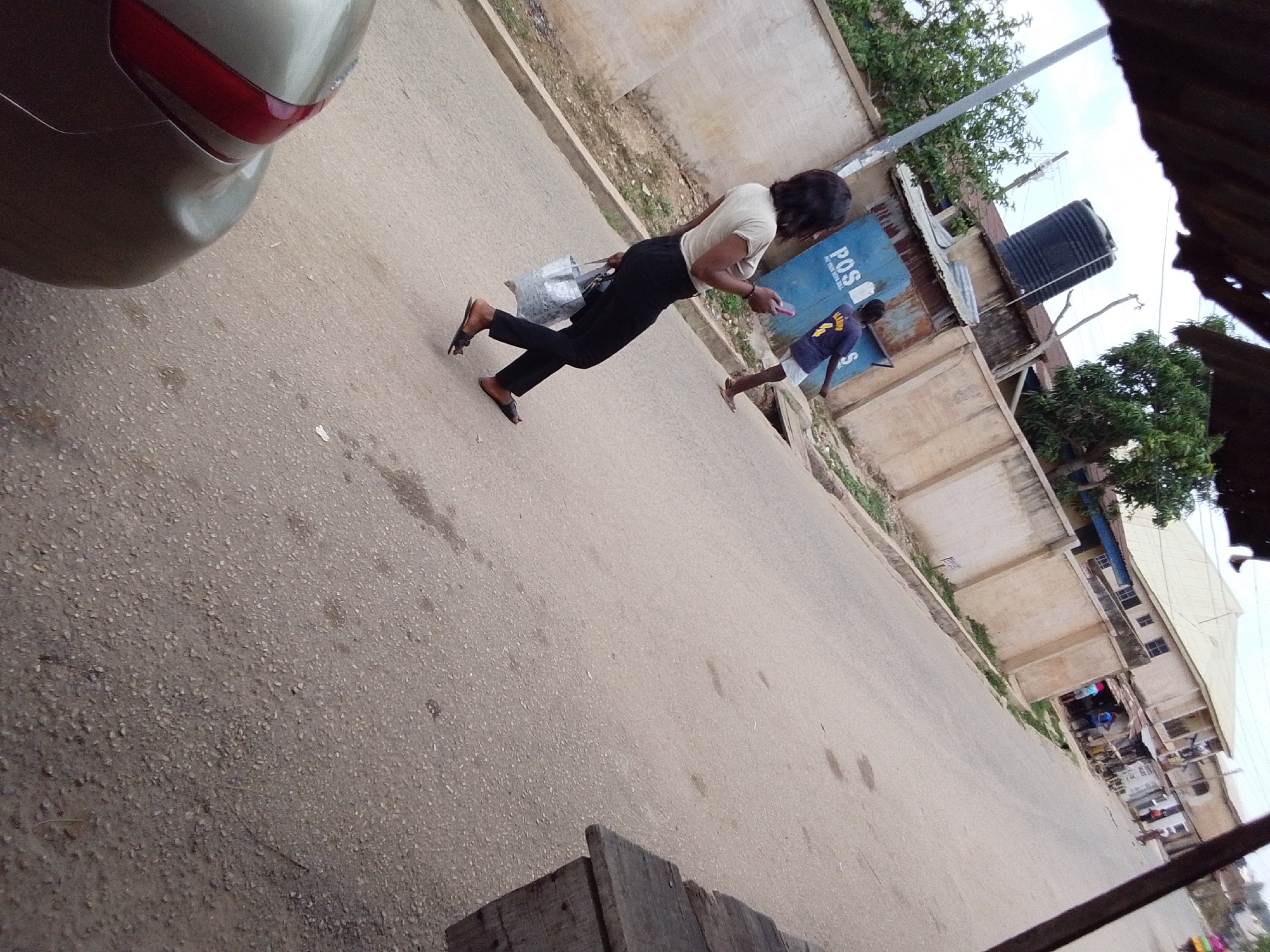 The image shows a street scene in Jos, Nigeria. A woman wearing a light-colored top and dark pants is walking on the side of the road, looking at a mobile phone. She carries a silver-colored tote bag. A young man wearing a blue shirt is walking a short distance behind her, also on the side of the road. The backdrop includes buildings with a blue P.O.S sign, a water tank, and some greenery. In the foreground, the rear of a vehicle is visible on the left side of the road. The road appears to be paved.