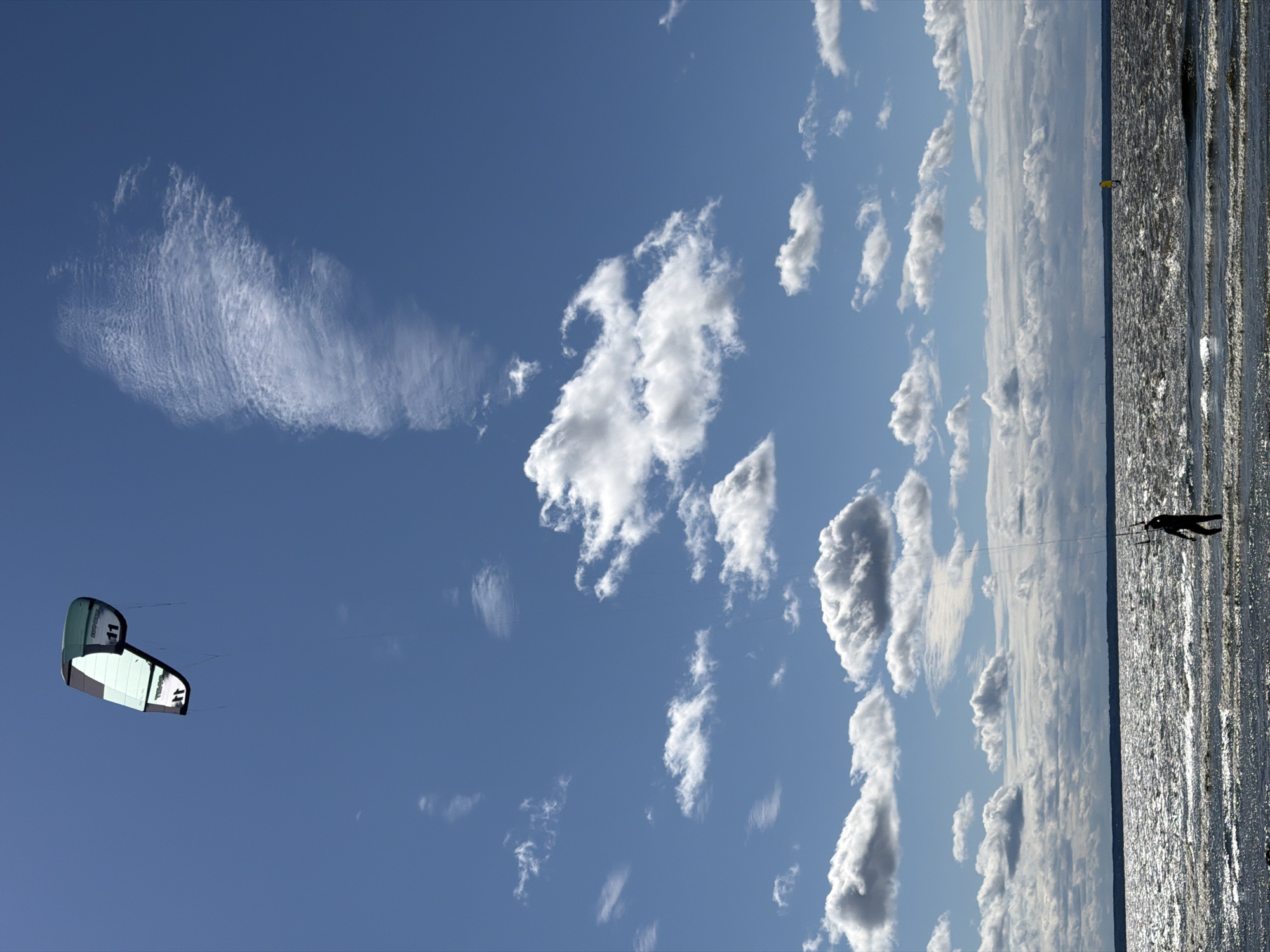 A kite surfer is seen in silhouette, standing in the shallow water of the Gulf of Finland. The surfer is holding onto the control bar, with the kite visible in the bright blue sky above. The kite is a vibrant green and white, with the number "11" clearly displayed on it. The water is choppy, reflecting the sunlight and creating a shimmering effect. The sky is dotted with scattered white clouds, and a wispy cirrus cloud stretches across the upper right portion of the frame. In the distance, a faint landmass is visible on the horizon, and a yellow marker buoy can be seen in the water. The scene suggests a windy day, ideal for kitesurfing in this coastal area near Kronstadt, Russia.