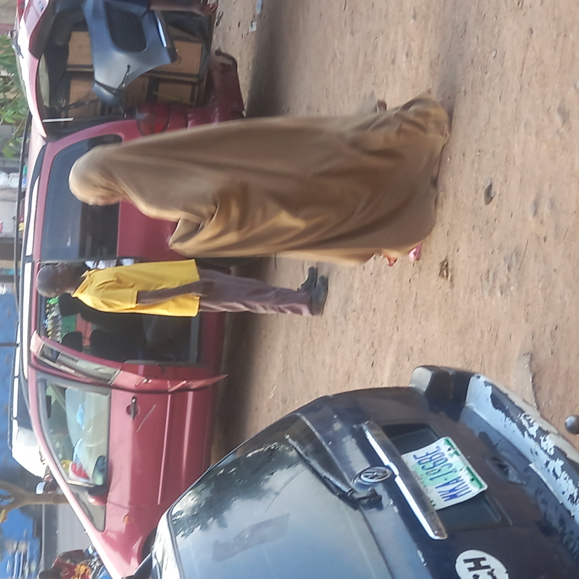 The image depicts an outdoor scene in what appears to be a street or roadside area in Dadi Riba, Nigeria. The ground is unpaved, consisting of dirt or sand.
Two people are visible. One person, to the left (when the image is correctly oriented), is a man wearing a bright yellow short-sleeved polo shirt, dark brown or grey trousers, and dark shoes. He is standing near the open door of a red van or minibus. The other person, positioned slightly behind and to the right of the man, is covered in a full-body, light brown or tan garment that obscures their figure and face, consistent with religious attire.
The prominent object in the midground is a red van or minibus, possibly a commercial passenger vehicle. Its rear is open, revealing several car parts, including what looks like a black car bumper and cardboard boxes, suggesting it might be undergoing repair, being loaded/unloaded, or used for transporting goods.
In the foreground, the hood and windshield of another vehicle, dark blue or black, are partially visible. On its hood, there is a Nigerian license plate clearly displaying the alphanumeric code "MKA 795BE" against a green and white background.
The lighting suggests it is daytime, possibly morning or afternoon, with bright and relatively even illumination. No specific activity beyond standing and observation can be definitively determined for the individuals.
